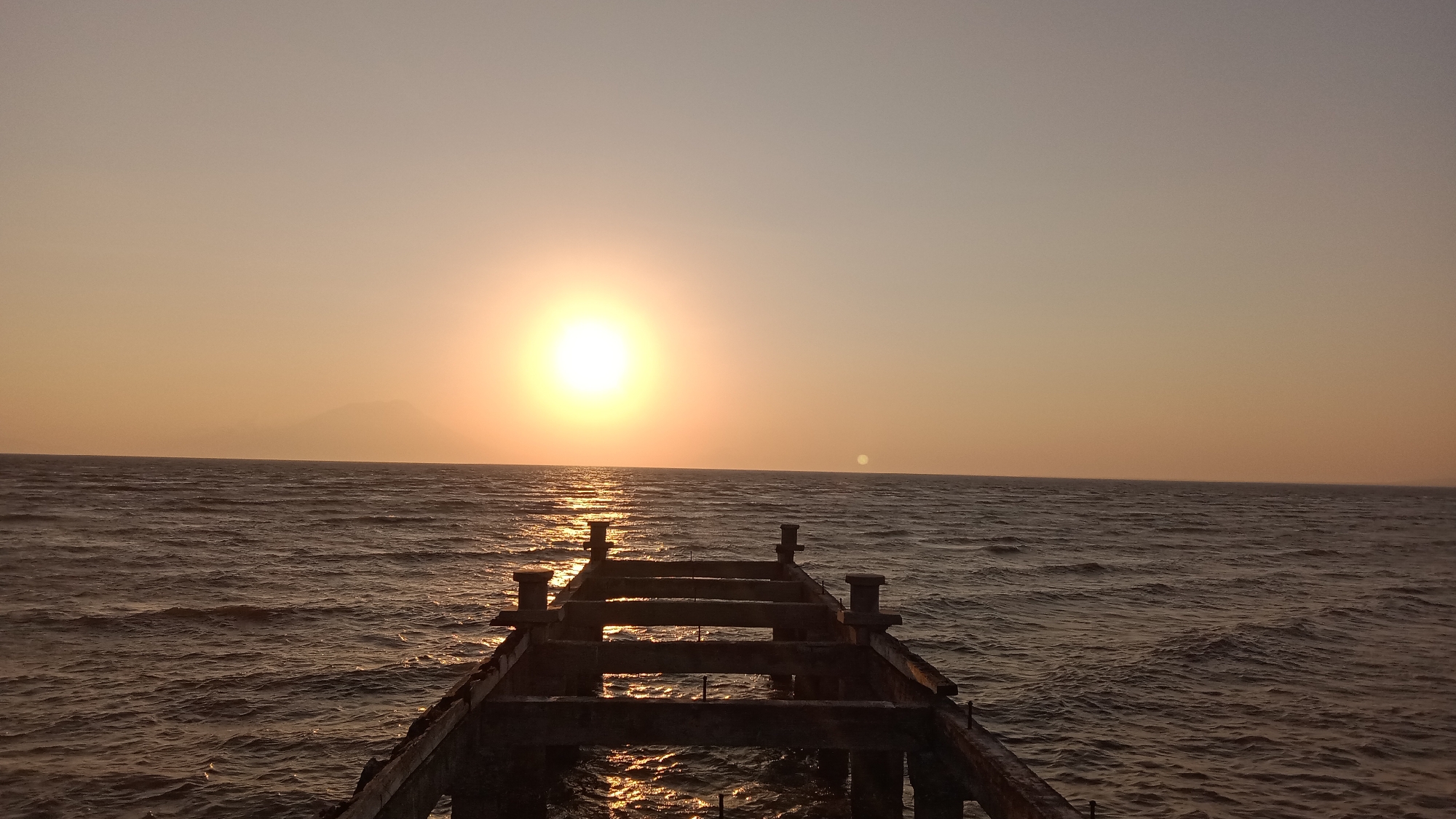 The image shows a sunset over the sea. The sun is a bright orb in the sky, casting a golden glow on the water. A derelict pier extends into the sea, its concrete structure silhouetted against the sunset. The water is choppy, with small waves creating a textured surface. In the distance, a hazy mountain range is visible on the horizon. The scene is peaceful and serene, with the natural beauty of the sunset and the sea dominating the composition. The location is not specified beyond "Unknown, Indonesia."