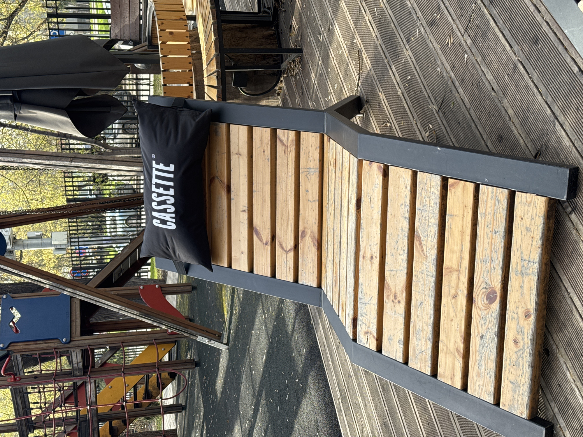 The image shows a wooden chaise lounge with a black pillow that says "CASSETTE" in white letters, located on a wooden deck next to a playground. The playground equipment is brightly colored with red, blue, and yellow elements. The scene appears to be outdoors, possibly in a park or recreation area. The city is Moscow, Russia. There are trees visible in the background.