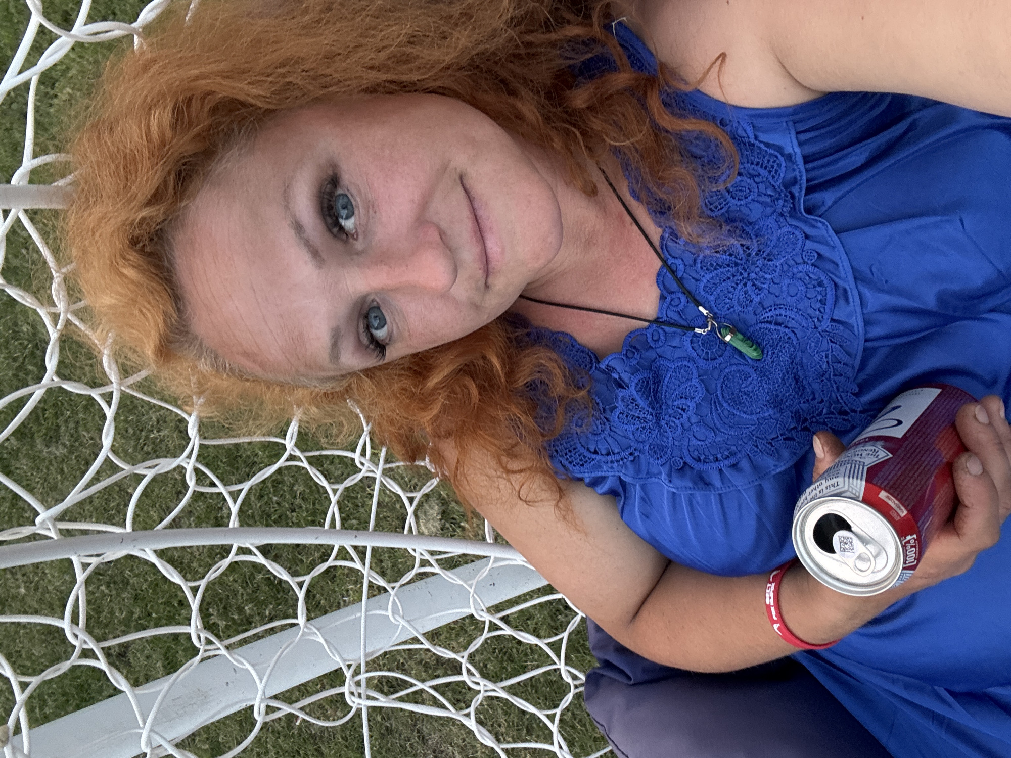 In the city of Chaikovsky, Russia, a woman with vibrant red, curly hair is seated on a white, net-like swing against a backdrop of lush green grass. She is wearing a bright blue dress with intricate lace detailing on the bodice and a dark pendant necklace with a green stone. In her right hand, she holds a red and silver can. A red bracelet adorns her left wrist. The scene appears to be outdoors, possibly in a garden or park setting.