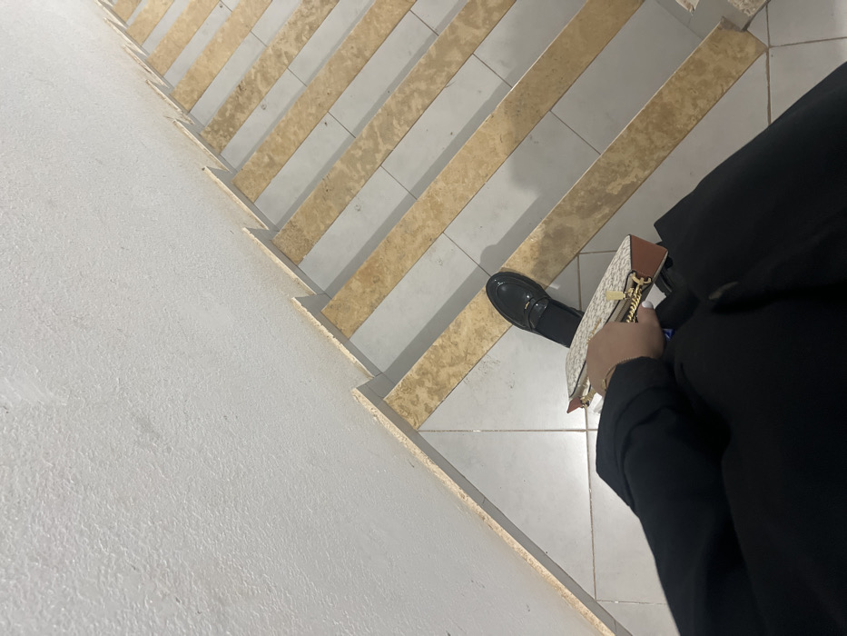 A person stands on a staircase. The steps feature alternating light beige wood-look planks and white tiled risers. The person wears a black outfit and black shoes. They hold a beige wallet with a brown trim and gold zipper. The camera angle is downward, looking from above. The staircase appears to be in a modern, indoor setting.