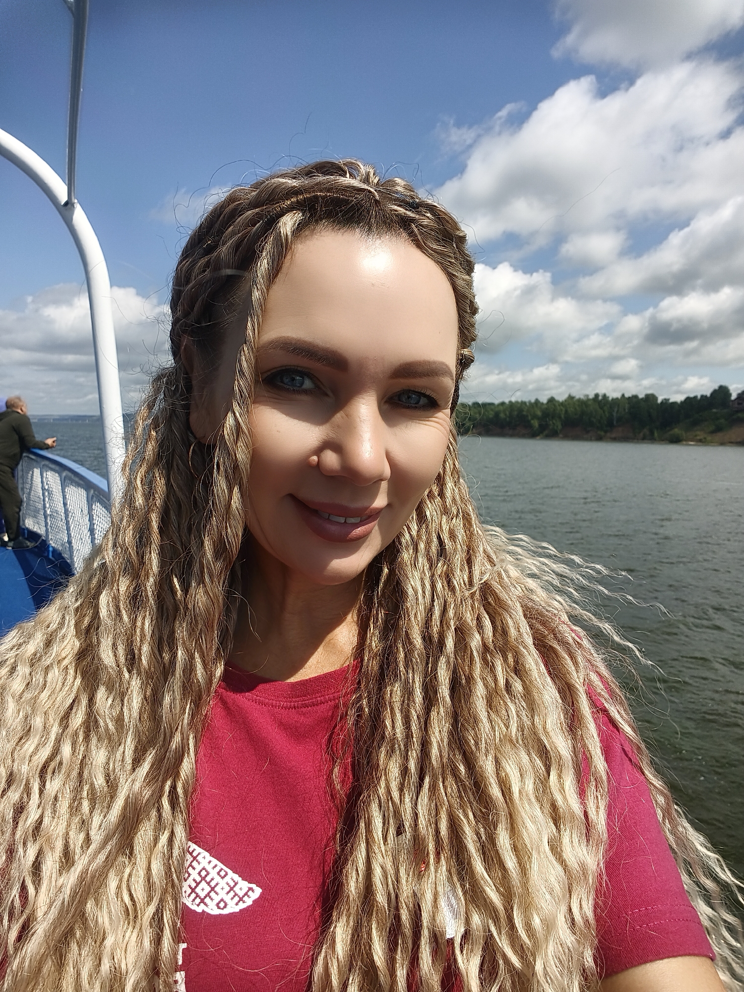 The photograph captures a close-up selfie of a woman with blonde, braided hair, taken from the chest up. She is wearing a red t-shirt with a white embroidered pattern on the left side of her chest. The woman is smiling and looking towards the camera.
The background reveals a body of water, likely a river, with a tree-lined shore visible in the distance. The sky is blue with scattered white clouds. To the left of the woman, a white railing and a portion of a white pole are visible, suggesting she is on a boat or a pier. A man in a dark outfit is standing further down the railing, looking out at the water.
The context provided specifies that the location is Shelanga, Russia. The scene appears to be a scenic outdoor setting on a sunny day.