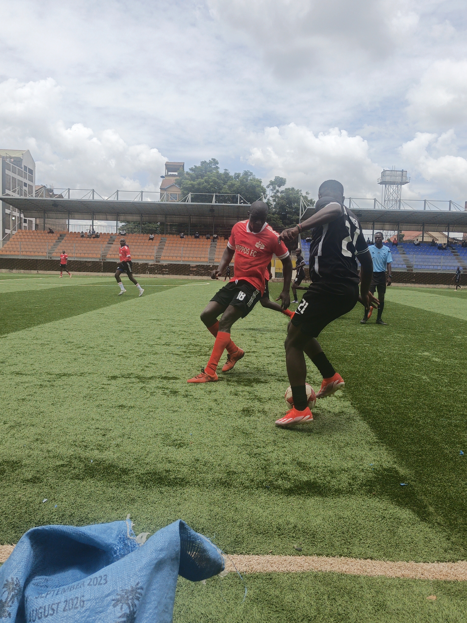 The photograph captures a soccer match in progress on a green artificial turf field. Two players are the central focus: one in a red jersey with "HIPPOS FC" on the front and the number 18 on the back, and the other in a black jersey with the number 21. The player in black is dribbling the soccer ball. Both wear matching-colored cleats.
In the background, other players are visible on the field and a referee is seen wearing a light blue uniform. The stadium's seating is a combination of orange and blue. In the far background, buildings can be seen, including a taller building and a water tower. A blue sack with the text "SEPTEMBER 2023" and "AUGUST 2026" is in the foreground. The sky is partly cloudy.
The stadium is likely located within Nairobi, Kenya, evidenced by the architectural style of the buildings and the overall environment.