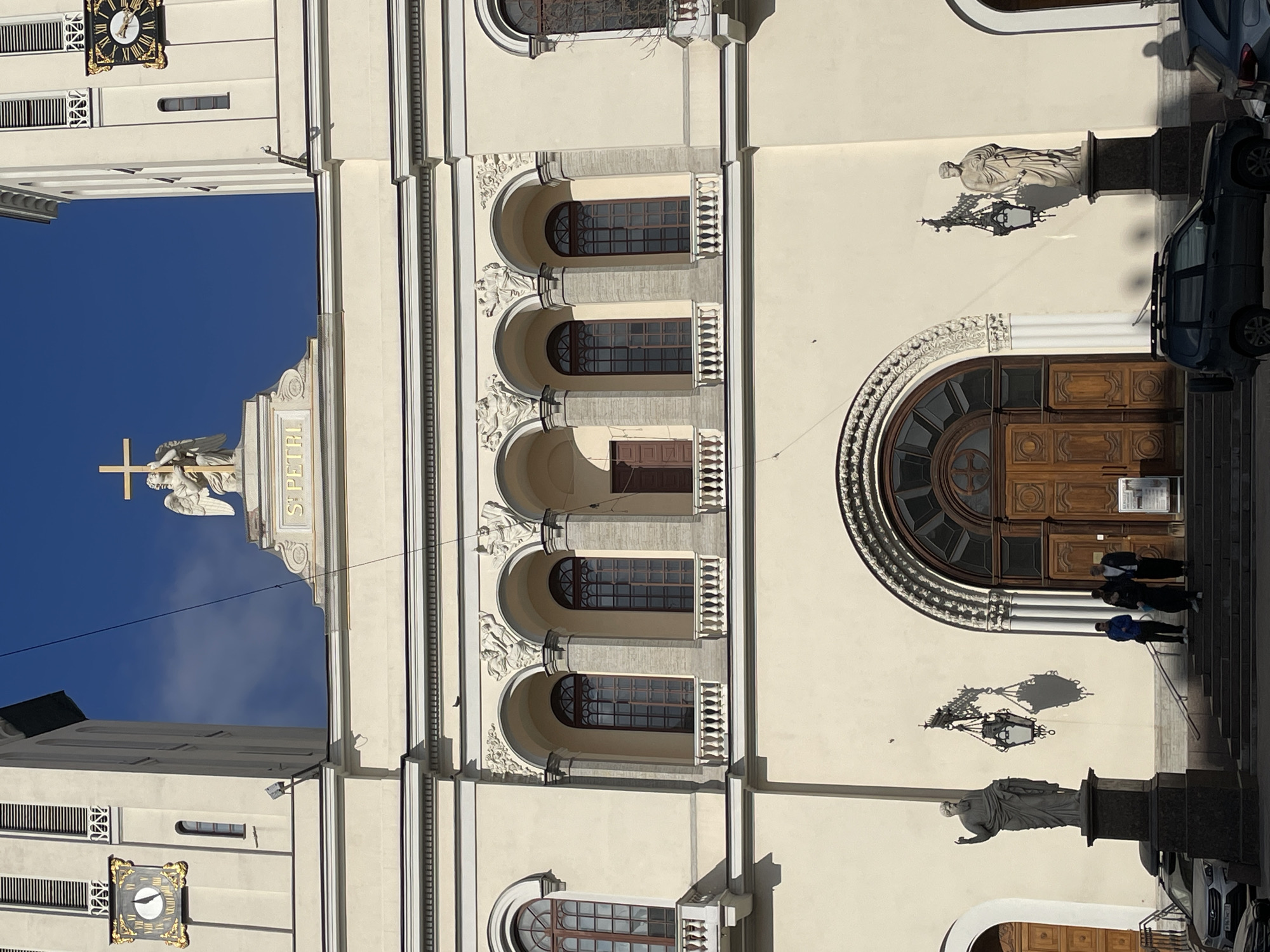 The image depicts the St. Peter and Paul Cathedral (St. Petri Kirche) located at 22-24 Nevsky Prospect in Saint Petersburg, Russia, under a clear blue sky. The building's facade is predominantly light cream. Two symmetrical bell towers flank a central pediment, where a white angel statue holds a golden cross above the inscription "S. PETRI." Each tower features a gilded clock face with Roman numerals; the left clock indicates approximately 9:00, and the right approximately 10:00.
Below the pediment, an upper level contains five arched openings. The central arch is recessed, while four flanking arches feature dark-framed windows, two of which have small balustrades. Ornate stucco decorations are present above these arches.
The ground level features a large, arched double-door main entrance constructed from dark wooden panels with intricate carvings. This entrance is accessed via a wide set of dark gray steps. Two classical-style statues on dark pedestals flank the steps; the left statue gestures with an upraised arm, and the right holds an object. Two ornate black metal lanterns are affixed to the facade, casting distinct shadows to their left.
Four individuals are present on the steps. Three are grouped near the main entrance, dressed in dark and blue casual attire. One individual stands further right, wearing a lighter jacket.
Two vehicles are parked on the street. A silver crossover SUV with a roof rack is on the left, displaying a Russian license plate (967 CT 178). A dark-colored SUV with a roof rack is partially visible on the right. Bright sunlight illuminates the scene, creating sharp shadows. Thin black wires traverse the upper portion of the frame. A small bird is perched on a facade ledge.