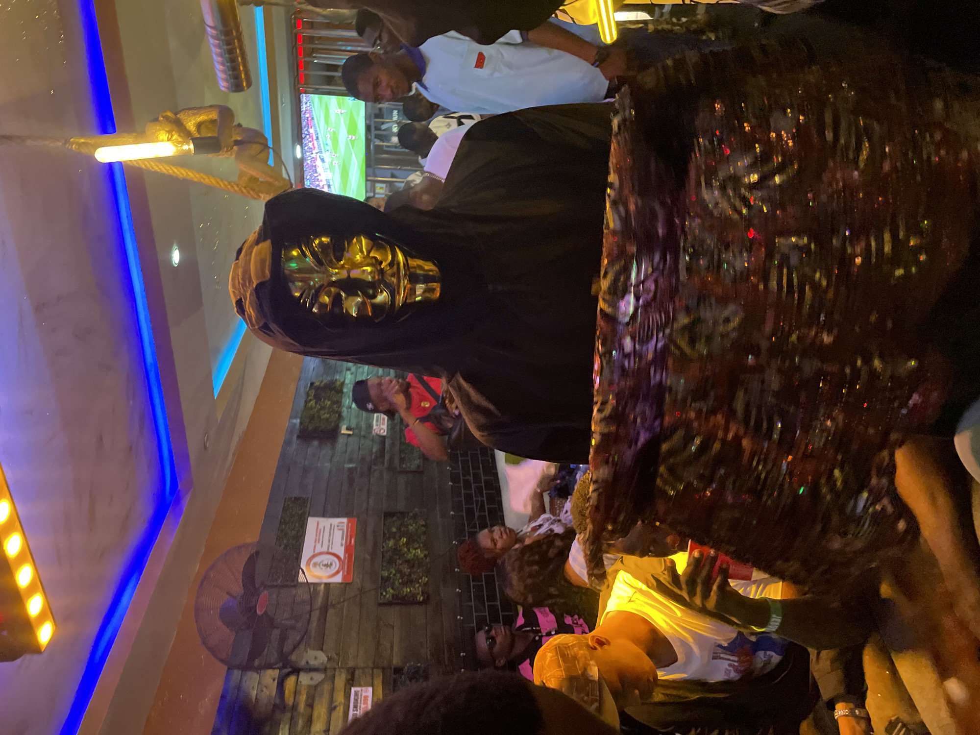 The image shows a crowded indoor space, likely a bar or club, in Obosi, Nigeria. A person wearing a black hooded cloak and a golden mask is prominently positioned in the foreground. In the background, several people are visible, some watching a soccer match on a television screen. Other details include wooden wall paneling, a ceiling fan, decorative lighting, and various individuals engaged in conversation and social interaction.