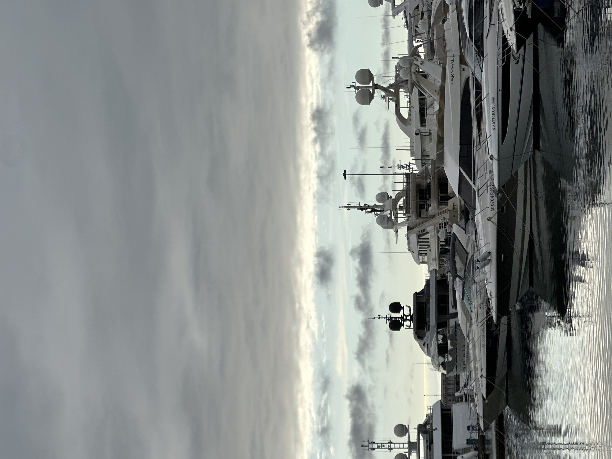 A marina in Sochi, Russia, is depicted under an overcast sky exhibiting varying shades of grey and intermittent lighter, bluer cloud patches. The foreground and midground feature numerous vessels moored in calm, dark water that reflects the boats and sky. Visible vessels include several white motor yachts, one marked "AC 0764 RUS 2" and another "XA0770RUS50" on their hulls. A larger yacht named "SKYFALL" is also present. Further back, multiple multi-decked superyachts, predominantly white and one with a grey hull, are docked, displaying intricate superstructures, radar domes, and communication masts. A utility boat bearing the name "TAURICA" is partially visible among the larger vessels. No individuals are present, and no active movements or interactions are observable. The scene is illuminated by diffused natural light.