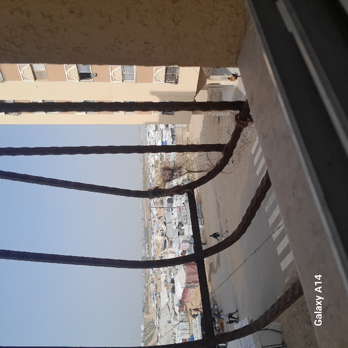 The image, taken from an elevated perspective in Khan Yunis, Palestinian Territory, depicts a scene encompassing both urban structures and an extensive informal settlement. The foreground is framed by four thick, dark, rusty vertical metal bars, slightly bowed, with some dried foliage clinging to one on the right, and a beige window sill with a horizontal metal frame at the bottom.
Immediately beyond this framing, a dirt-colored road with a white zebra-stripe pedestrian crossing is visible. On this road, multiple individuals are present: two figures are grouped on the left, one in dark attire, the other in lighter clothing; a lone figure in a dark shirt and light trousers walks rightward, appearing to kick something on the ground near the center. To the far right, a person in a yellow top and dark trousers sits near the base of a multi-story, light-beige concrete building that occupies the right side of the frame. This building features multiple rectangular windows, some with dark grilles and decorative arch patterns in a lighter hue.
The background reveals a vast, densely packed informal settlement extending across the landscape, composed of numerous white and light-colored tents and makeshift shelters constructed from tarpaulins and other materials. Beyond the settlement, sandy, undulating terrain is visible on the distant horizon to the left. The sky overhead is clear and light blue, indicating daylight conditions. The watermark "Galaxy A14" is present in the lower-left corner.