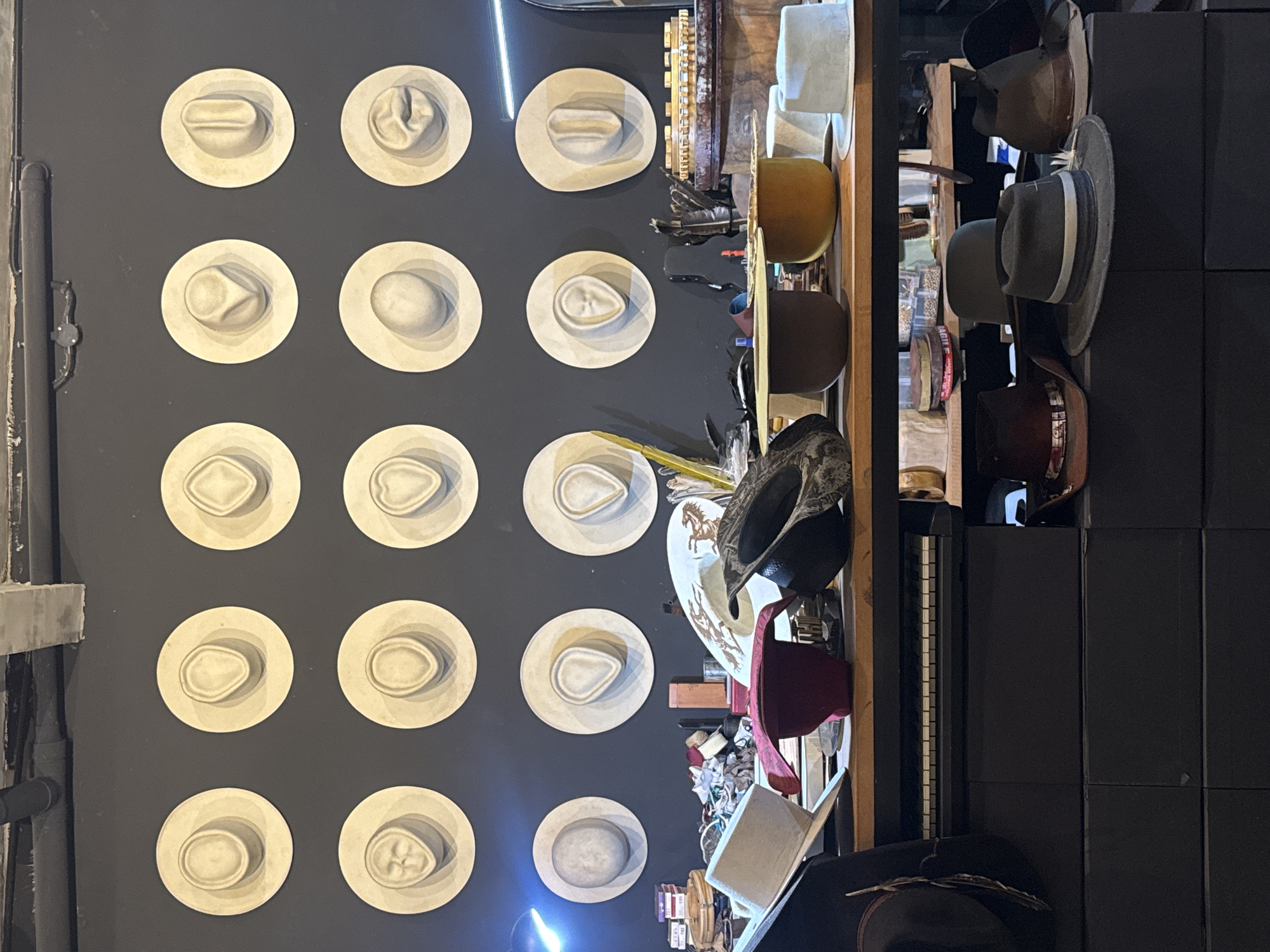 The image depicts the interior of a store in North Kuta, Indonesia, likely a hat shop. The left wall is dark gray, adorned with twelve circular displays, each showcasing a molded, white-painted hat form. To the right of this display, a shelf is stocked with a variety of hats in different styles and colors, including cowboy hats, fedoras, and wide-brimmed hats. Other objects, such as tools and accessories, are also visible on the shelf. The right side of the image shows a wall with dark square tiles.