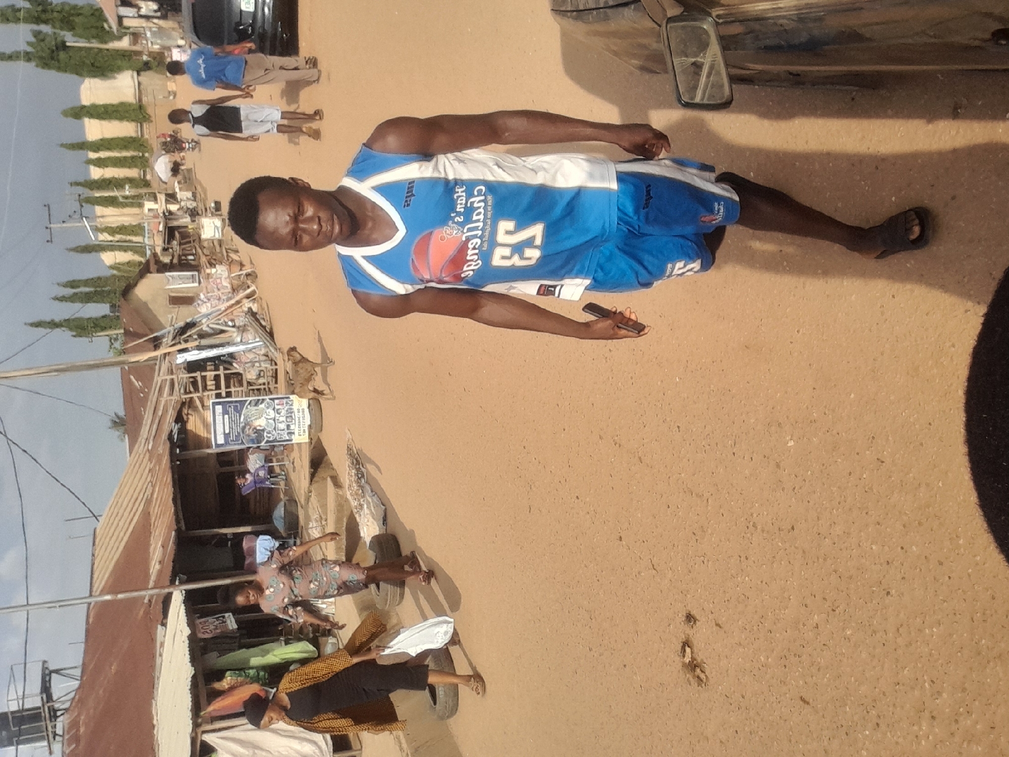 In a dusty, unpaved street in Abuja, Nigeria, a young Black male walks towards the viewer, wearing a blue and white basketball jersey with the number 23 and matching blue shorts. He holds a dark mobile phone in his right hand. To his left, two women walk away from the viewer. The woman in front wears a patterned dress, and the woman behind her wears a black dress and a patterned yellow and black scarf. She carries a white shopping bag in her left hand. In the background, several small, weathered structures with corrugated iron roofs line the street. Several utility poles and wires are visible against a hazy sky. A brown dog is visible on the right side of the street, further down. A black car is parked on the right side of the image, partially obscuring the view of the street beyond. The ground is a light brown, sandy soil with scattered debris.