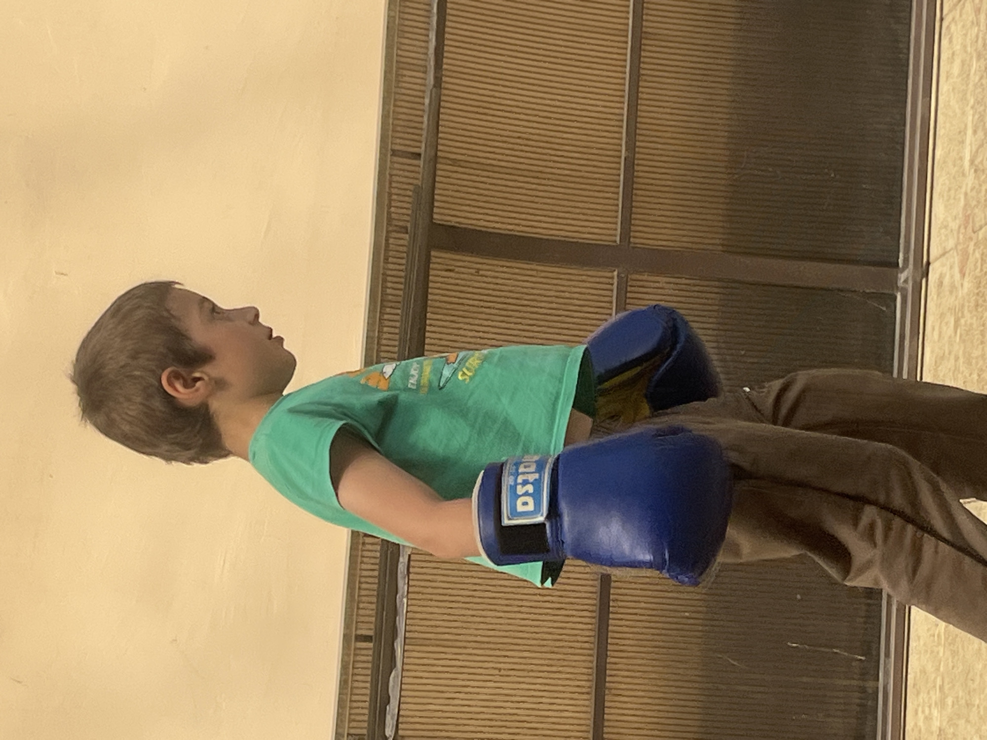 An East Asian boy, with brown hair and fair skin, stands facing right, wearing a green t-shirt and brown sweatpants. He has blue boxing gloves on both hands. The boy appears to be in a boxing stance, with his head lowered and looking down. The background consists of a ribbed, brown wall and a light yellow wall. The scene is indoors, and the lighting is moderate. The image appears to have been taken at an unknown location in the city of Kaspiysk, Russia.