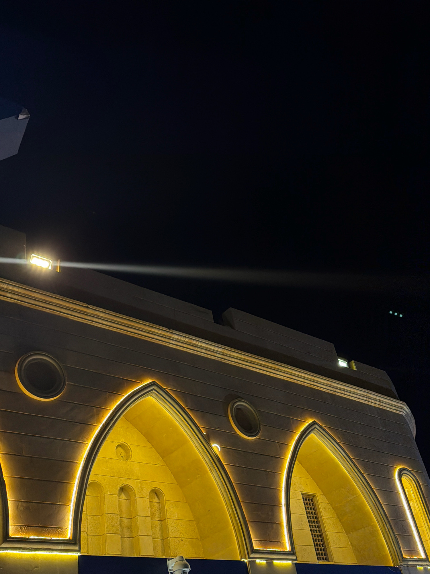 The image shows the illuminated facade of a building at night in Sharm El Sheikh, Egypt. The building has arched openings, with yellow lights outlining the arches. There are also circular windows on the upper part of the facade. A bright light is visible on the roof, casting a beam across the dark sky. In the distance, three small green lights are visible in the sky, possibly an aircraft. The overall impression is one of an illuminated architectural feature against a dark night sky.