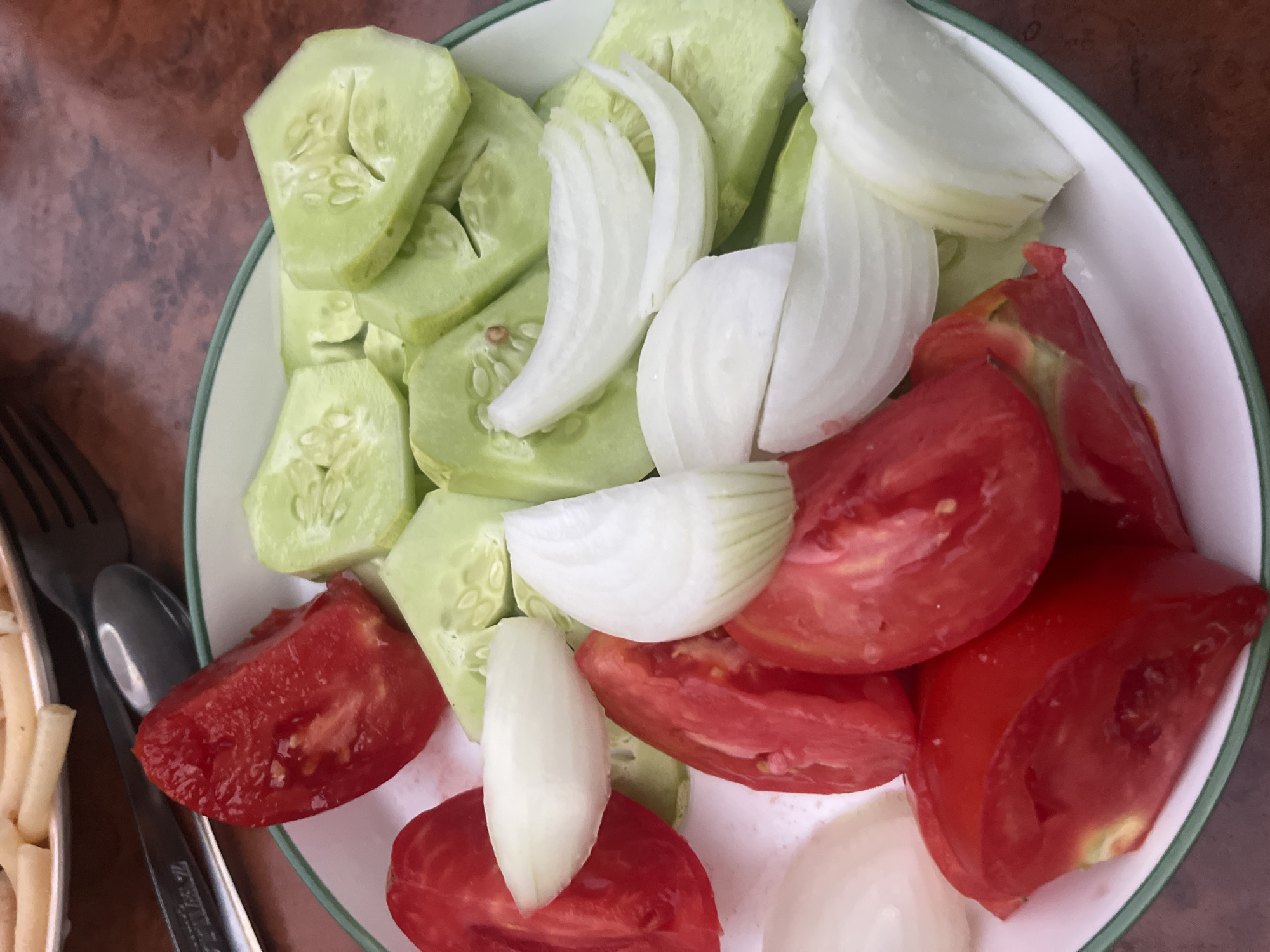 A close-up, overhead shot shows a white plate with a green rim, filled with sliced tomatoes, cucumbers, and white onions. The vegetables are artfully arranged on the plate. In the upper left background, a portion of another dish, possibly pasta, is visible, along with a fork and spoon. The plate rests on a wooden surface. The lighting is natural, casting soft shadows. There are no people visible in the image. The setting appears to be a mealtime scene. No specific location in Muhametjan Tüymebaev, Kazakhstan, is identifiable from the image.