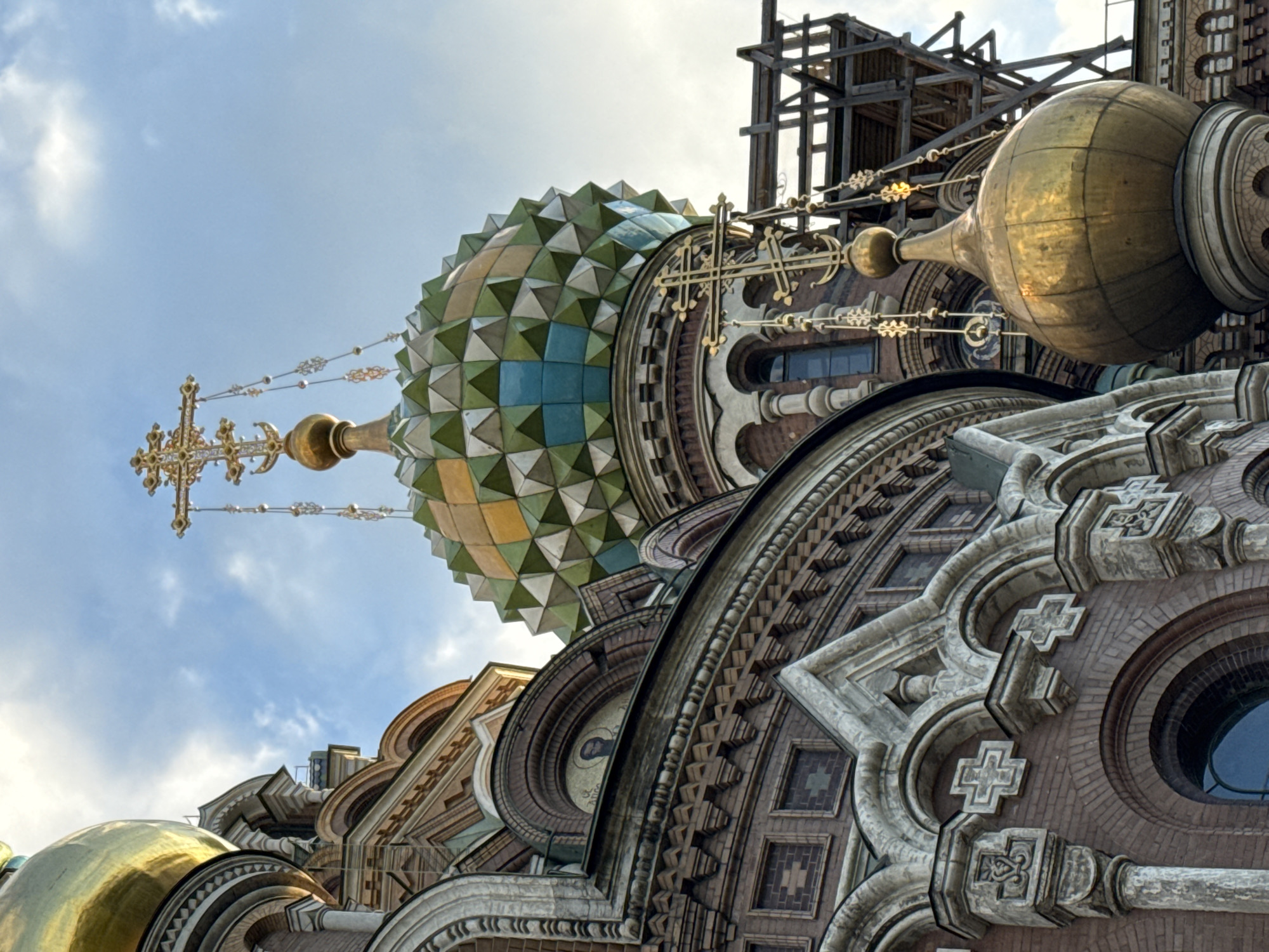 This is a medium-angle, eye-level, slightly low shot of the Church of the Savior on Spilled Blood in Saint Petersburg, Russia. The sky is partly cloudy and a light blue. The foreground is dominated by the ornate architecture of the church, with a prominent golden dome on the right. Several other domes and architectural elements are visible, including a large green and yellow faceted dome topped with a gold cross. The facade is constructed of red brick with intricate decorative stonework and mosaic elements. Scaffolding is visible on the right side of the image, obscuring some of the structure. No people are present in the image.