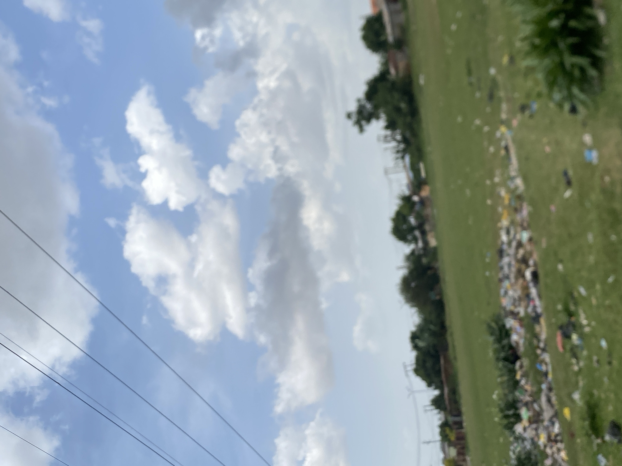 The image shows a view in Bukuru, Nigeria, with a blue sky partially covered by white and grey clouds. Electrical power lines stretch across the sky in the lower left of the frame. A green field, heavily littered with trash, extends to the right. Scattered trees and buildings are visible at the edge of the field. The image appears to be slightly blurry.