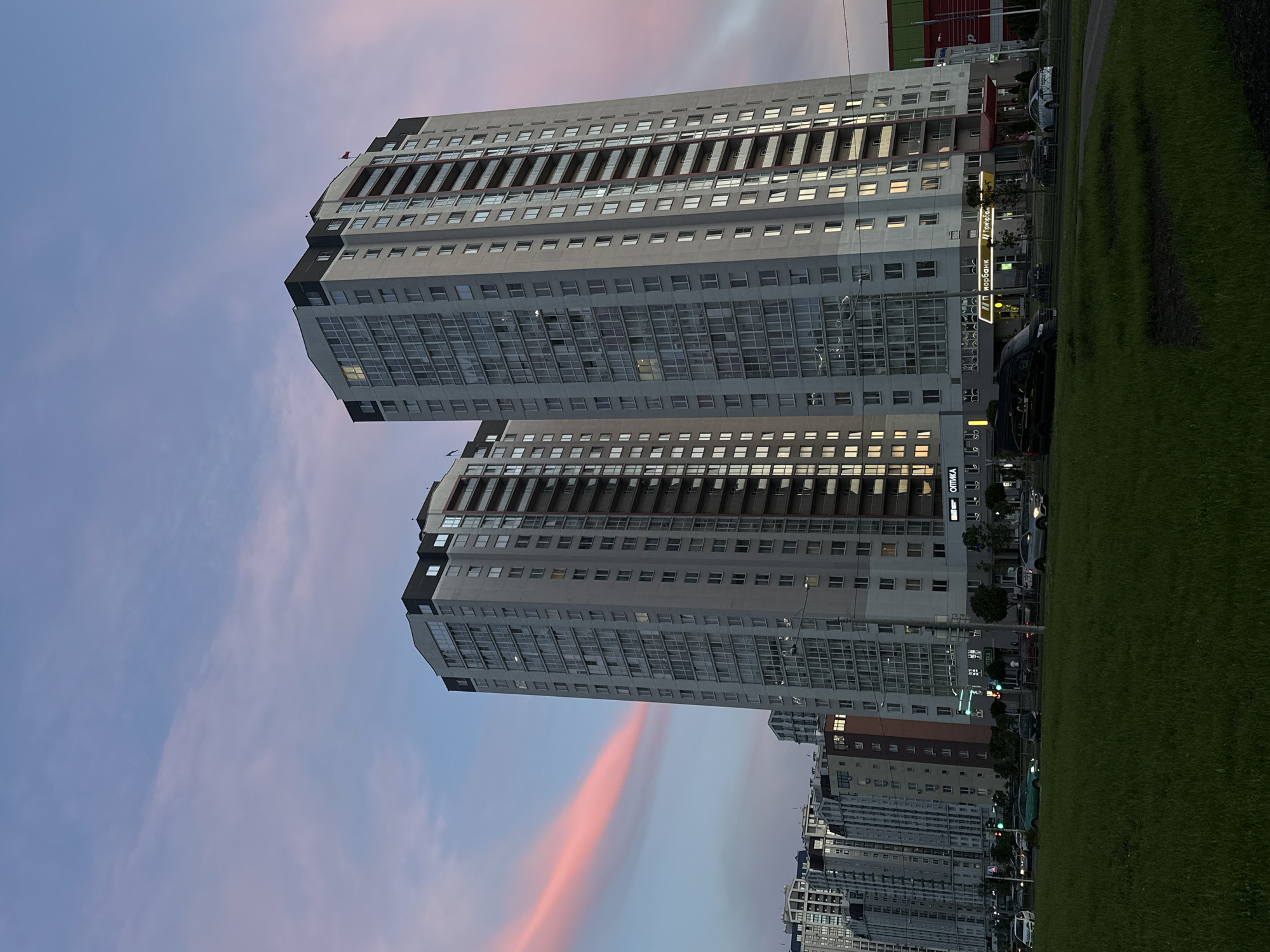 This image captures an urban landscape at either dawn or dusk in Minsk, Belarus, characterized by modern high-rise residential buildings and a prominent green space. The two most prominent buildings stand tall on the left side of the frame, featuring multi-story facades with numerous windows, balconies, and a mix of grey, dark, and glass elements in their architecture. Lights are visible in some of the windows, indicating the time of day. More buildings extend into the distance at the lower right.
The sky, occupying the upper portion of the scene, displays soft hues of blue, pink, and orange, with distinct elongated clouds streaking across, particularly a vivid pink cloud closer to the horizon, reinforcing the impression of a beautiful sunrise or sunset. The right side of the image is dominated by a neatly maintained green lawn, suggesting a park or public area, bordered by a darker path or ground covering.
Along the base of the buildings, several parked cars are visible, alongside trees and streetlights, suggesting a street or access road. A brightly lit sign on the ground floor of one building clearly displays the Cyrillic text "Приорбанк" (Priorbank), indicating a financial institution. Another sign further down, partially obscured but visible in crops, reads "ОПТИКА" (Optics). No people are distinctly visible, contributing to a calm and tranquil atmosphere. The scene highlights contemporary urban development set against a striking sky.