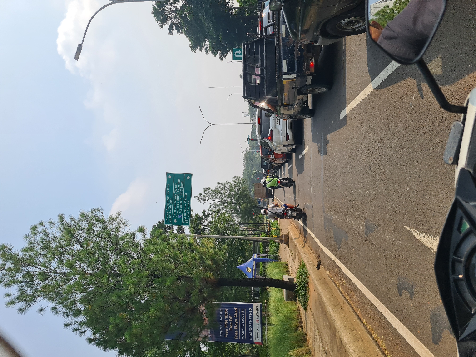 An image captures a busy daytime street scene from the perspective of a motorcycle, with a portion of the motorcycle's mirror and fairing visible in the foreground. The road is experiencing traffic congestion, featuring multiple cars, including a black pick-up truck and a white car with the license plate B 1127. Several motorcyclists are seen navigating the road, one in a green jacket and another in a white helmet.
The sky is bright with scattered clouds, indicating sunny weather. A tall streetlight arches over the road. To the left, a large evergreen tree dominates the midground, with green grass and foliage lining the roadside. Further back on the left, a blue tent is set up, and a large green road sign provides directional information.
The text on the road sign includes destinations such as "Foresta, Nava Park, De Park," "Damai Indah Golf," "Central Business District," "Convention (ICE), Aeon Mall," "Tol Jakarta - BSD Barat," "East Business District," and "RS. Eka Hospital." Below the tree, a banner advertises "SOHO SIGNATURE" with details like "Free PPN 100%", "Free DP", "Free Biaya Akad", and "READY TO MOVE IN!". A contact number, "0813-771-771-90", is also visible, along with company names "DHIJAYA KARYA", "sinarmas land", and "MA BANGUN SENTANA". The scene suggests an urban or suburban area, likely in Indonesia, given the local context of the named locations and vehicle plate.