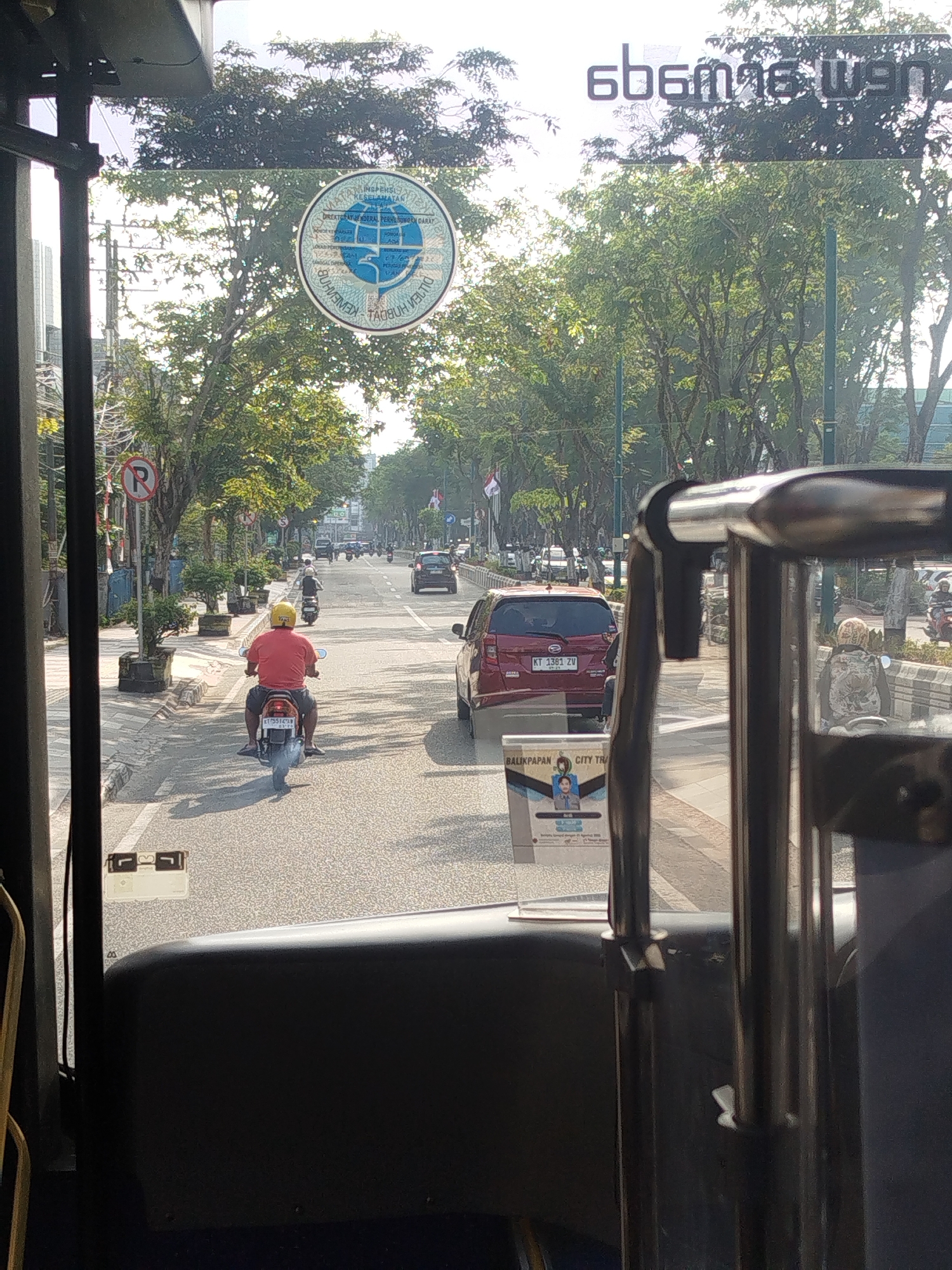 This is a first-person view from inside a vehicle, looking out through the windshield at a city street scene in Balikpapan, Indonesia. The foreground shows the interior of the vehicle, including a dark-colored dashboard and metal bars. Outside, the street is lined with trees and buildings. Traffic is visible, including a red car and several motorcycles.
In the middle ground, a person wearing a red shirt and a yellow helmet is riding a motorcycle. The motorcycle has a license plate that reads "KT 5516 CA". The person appears to be heading away from the viewer.
On the left side of the street, a "P" parking restriction sign is visible. A circular emblem with text and a globe graphic is affixed to the windshield. On the right side of the windshield, the text "6b9n9g wen" is displayed in white lettering.
The overall lighting suggests it is daytime, possibly late morning or early afternoon, with sunlight filtering through the trees. The environment appears to be urban, with paved roads, sidewalks, and streetlights. The image captures a moment of everyday life in Balikpapan.