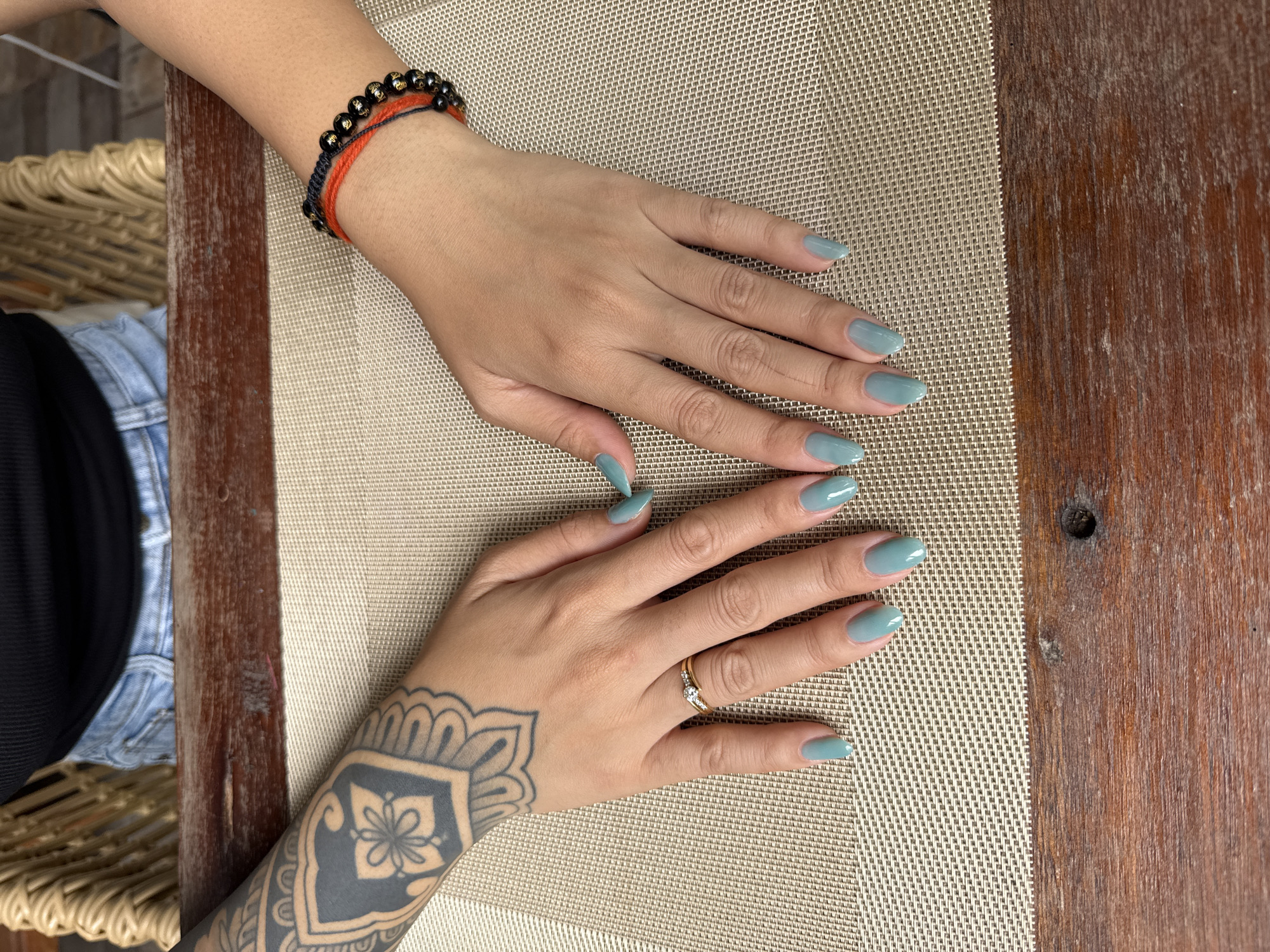 A high-angle medium shot shows a person's hands resting on a light brown woven placemat on a wooden table. The person has a large black tattoo on their left forearm and is wearing a black tank top and denim shorts. On their left hand, they are wearing a gold ring with small diamonds on the ring finger. Both hands have fingernails painted a light blue-green color. On the right wrist, there is a bracelet made of black beads and red cord. The background shows a woven chair and a brown wooden railing. The image appears to have been taken outdoors, possibly at a cafe or restaurant in Sai Ma Subdistrict, Thailand.