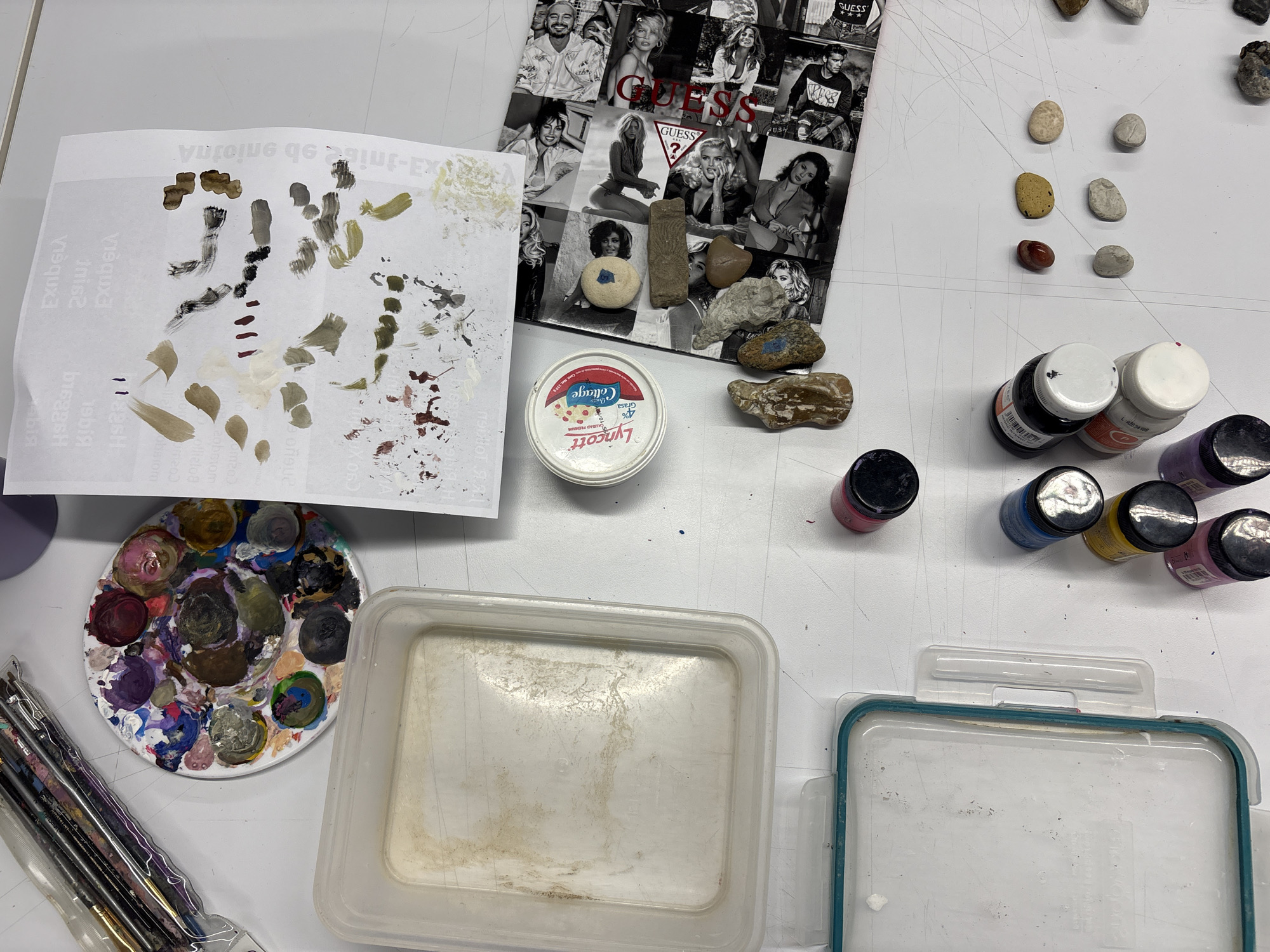 On a white surface, likely a table, an assortment of art supplies and objects are arranged. There are paintbrushes in a clear plastic case, a round paint palette with various colors, and several jars of paint with black lids, including one that is pink. There are two clear plastic containers, one larger and dirtier than the other, positioned near the paints. In addition to the art supplies, there is a white container of "Lyncott" cottage cheese, a black and white collage featuring Guess advertisements, and a collection of small stones. Some of the stones are adjacent to the collage, while others are grouped separately. Also present is a sheet of paper with paint swatches.
The setting is possibly an art studio or workspace in Mexico City, Mexico, based on the text in Spanish on the container of "Lyncott" cottage cheese. No people or actions are directly visible in the scene.