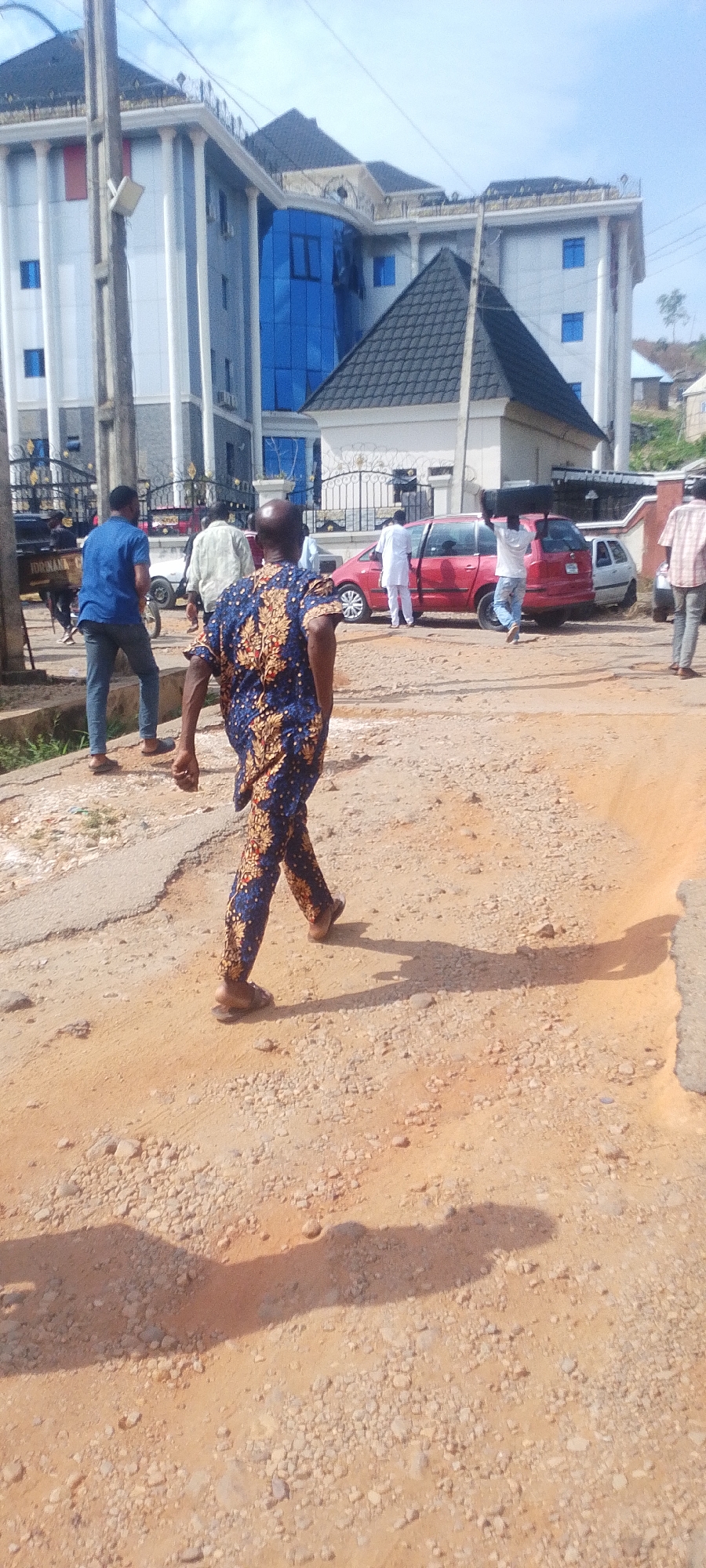 The image shows a group of people on a dirt road in Lokoja, Nigeria. A man in a blue patterned shirt and pants walks towards the foreground. In the background, a red van and a white car are parked near a modern building with blue tinted windows. Several other people are milling about. The environment appears to be a neighborhood or commercial area in the city.