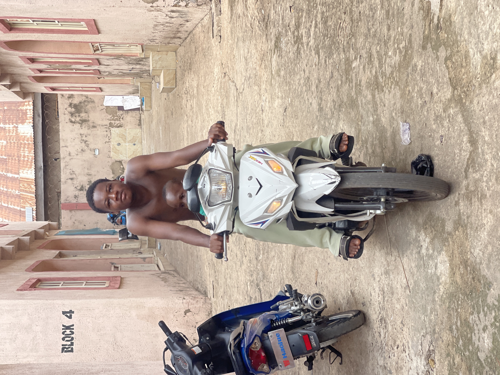 The image shows a shirtless man on a white motorcycle in an outdoor alleyway next to a building labeled "BLOCK 4," likely in Lokoja, Nigeria. Another blue motorcycle is nearby on the ground, displaying a "Haojue" license plate. The environment appears to be urban, possibly residential, and the man is looking directly at the camera. There is another person on the seat behind the shirtless man, but their details are obscured. The alley is paved and has litter visible. Clothes are hung to dry on the side of the building.