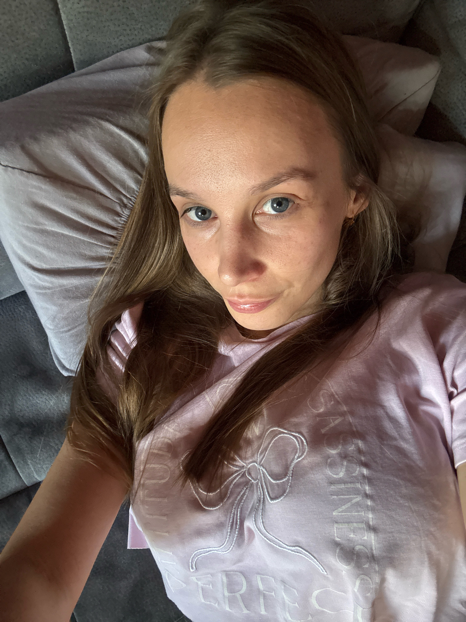 A selfie taken from a high angle shows a young woman with long brown hair and blue eyes lying on a gray sofa with a light pink pillow under her head. She is wearing a pale pink t-shirt with white embroidery, including a bow and text that appears to read "ASSINNESS" and "FREE". The lighting suggests an indoor setting, possibly a bedroom or living room. The image does not contain any identifiable elements that would place it specifically in Überherrn, Germany.