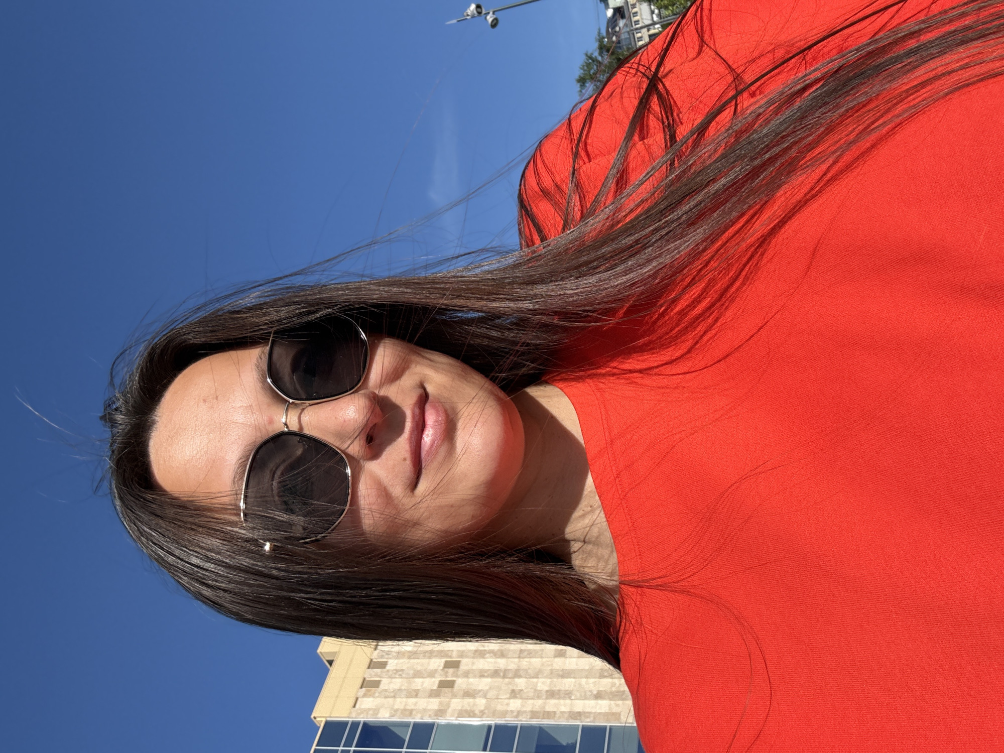 A woman with long dark hair and sunglasses is taking a selfie outdoors on a sunny day. She is wearing a red top. In the background, there is a clear blue sky and a building with glass windows. There is also a street light visible. The location is the city of Perm, Russia.