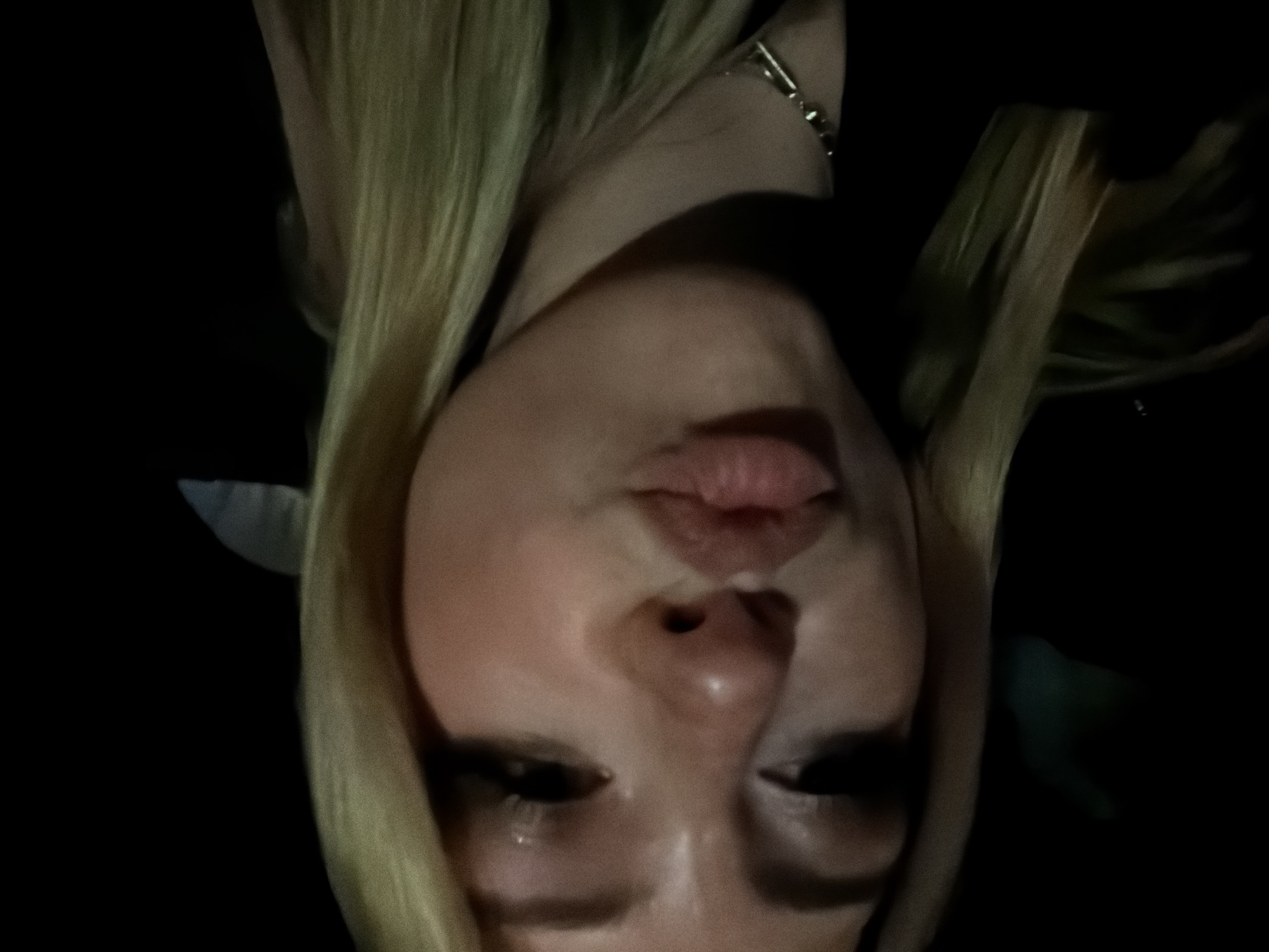 The image presents a close-up, head-and-shoulders portrait of a light-skinned individual, identified as a young female, situated in a low-light environment within the city of Paris, France. The subject is directly facing the camera with a puckered-lip expression, indicative of a "kissing" gesture. Her blonde hair is visible, framing the face, and a thin, silver-toned chain necklace rests against a dark garment. The eyes exhibit dark eyeliner and mascara, with a subtle shimmer on the eyelids. The background is predominantly black, devoid of discernible features, suggesting an indoor location or an outdoor setting at night with no ambient light sources providing specific context. Lighting on the subject is direct but soft, highlighting facial contours and makeup.
