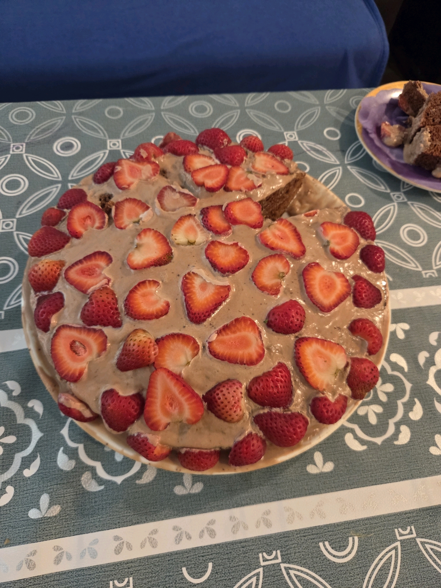 A chocolate cake, topped with a thick, beige frosting and slices of fresh strawberries, sits on a white plate. The cake is round and appears to be layered. The plate is positioned on a teal tablecloth with a white, geometric pattern. To the right of the cake, a small purple plate holds two slices of a dark-colored cake. The background is blurred, but a person wearing a blue shirt is visible. The setting appears to be a domestic environment, possibly a kitchen or dining room, in Glendale, United States of America.