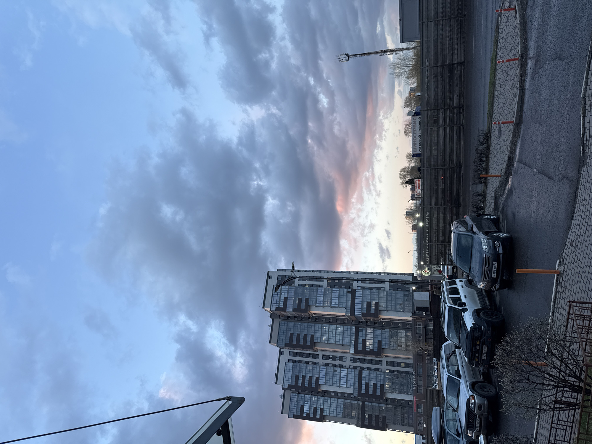 The image shows a street scene in Krasnoyarsk, Russia, at twilight. A tall, modern building dominates the left side of the frame, reflecting the sky in its many windows. Several cars are parked along the street, with their rear license plates visible. A metal fence and some utility infrastructure are visible in the background. The sky is filled with dramatic clouds, tinged with pink and orange from the setting sun. The overall atmosphere is urban and slightly muted due to the fading light.