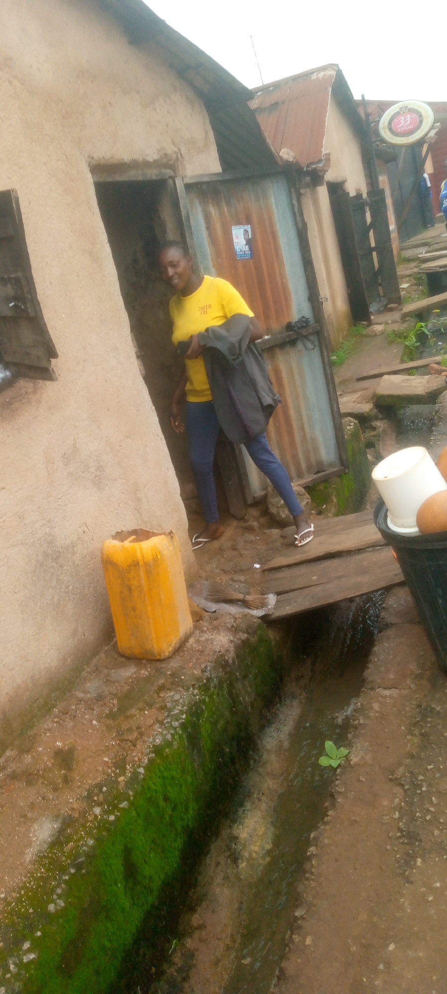 A person wearing a yellow t-shirt and blue pants stands near a doorway in an outdoor environment. The person is holding a dark jacket over their arm and appears to be smiling. The doorway leads into a building with a corrugated metal door. To the left of the doorway, a large yellow plastic container sits on the ground. In front of the doorway, a wooden plank bridge spans a narrow, shallow channel of water. The ground is dirt and some areas are covered with green moss. In the background, a sign with "33 EXPORT" is visible. The scene appears to be in a somewhat dilapidated urban setting.