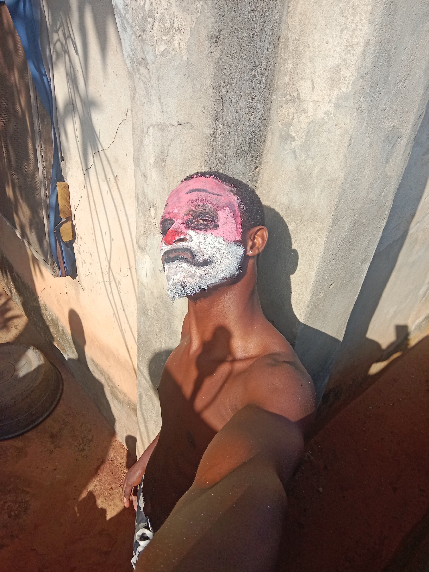 A shirtless man with pink and white makeup on his face stands outdoors in bright sunlight. The makeup resembles a clown's face, with a pink forehead and cheeks, a red nose, and a white beard and mustache. He is facing the camera, taking a selfie. He is located outdoors in Nkpor, Nigeria. The background is a textured concrete wall. A laundry line with clothes hanging on it is visible in the upper left corner. A metal wash basin is on the ground to the left of the man. The ground appears to be dusty or sandy. Harsh shadows are cast across the scene.