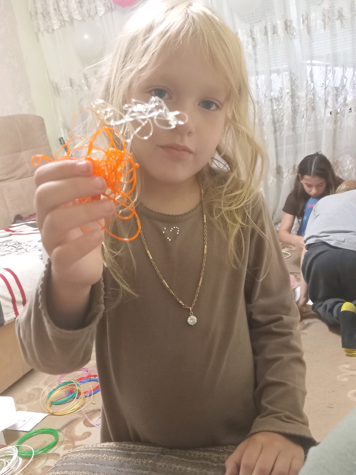 In Chuhuiv, Ukraine, a blonde-haired girl, wearing a brown long-sleeved shirt and a necklace with a pendant, holds up a tangled orange plastic filament from a 3D pen. She is looking towards the camera, and behind her, another girl and a person wearing a gray shirt and black pants are visible, along with spools of colored plastic filament on the floor. The setting appears to be an indoor space with lace curtains in the background and a patterned carpet.