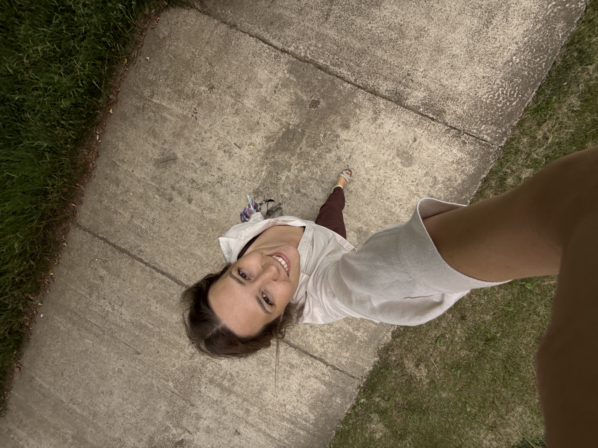 The image shows a woman taking a selfie outdoors. She is standing on a concrete sidewalk next to a grassy area. The woman is looking up at the camera and smiling. She is wearing a light-colored shirt and dark pants. Her feet are visible, wearing sandals. A bag with a colorful strap is hanging from her left shoulder. The lighting suggests it is daytime, possibly late afternoon or early evening, with soft shadows. The environment appears to be a suburban or park setting. The woman's expression conveys a sense of happiness and ease. There is no visible text in the image. The provided location context is Chaikovsky, Russia.