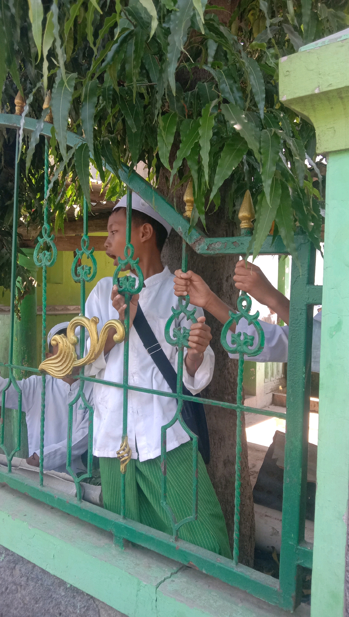 The image shows two young boys, likely students, behind a green metal fence. The boy in the foreground is wearing a white cap, a white tunic, and green striped pants. He is leaning forward and appears to be speaking or calling out, with his mouth open. His hands are gripping the fence. Another boy, mostly obscured by the fence and foliage, is visible behind and to the left of the first boy. His head is also covered with a white cap and he is wearing a white tunic. A third person's hands are visible on the fence to the right of the foreground boy, suggesting interaction. The fence is ornate with green scrollwork and gold decorative elements. Lush green foliage from a tree hangs over the fence and partially obscures the scene. In the background, parts of a building, possibly a mosque or madrasah given the attire of the boys, are visible. The ground appears to be made of concrete or stone. The overall lighting suggests a bright, sunny day. The location is identified as the Menara Kudus Mosque complex in Karang Anyar, Indonesia, a religious site characterized by its distinctive architecture and historical significance.