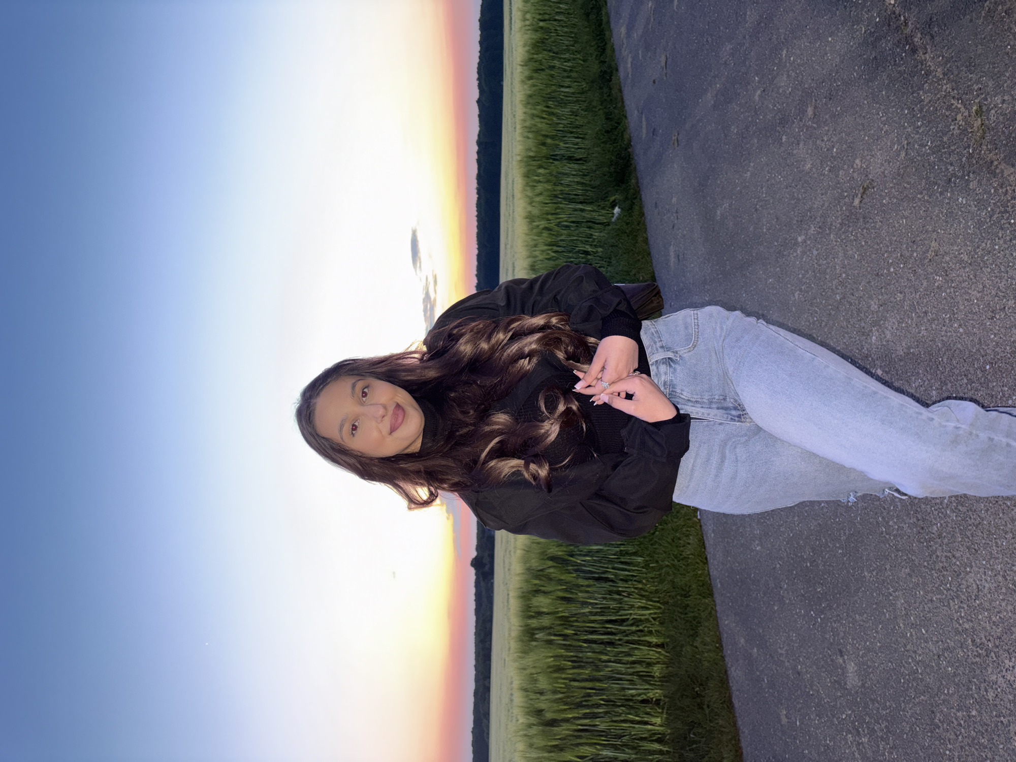 A young woman with long, brown, wavy hair is posed outdoors against a backdrop of a sunset sky and a field of green crops. She is wearing a black jacket and light blue jeans. The setting appears to be a rural area, likely near a road or path. The sky is gradient, transitioning from blue to orange and pink near the horizon. The photograph's angle suggests it was taken from a slightly lower viewpoint, and the landscape's angle might indicate a slightly rotated or angled shot. The location is Wegberg, Germany.