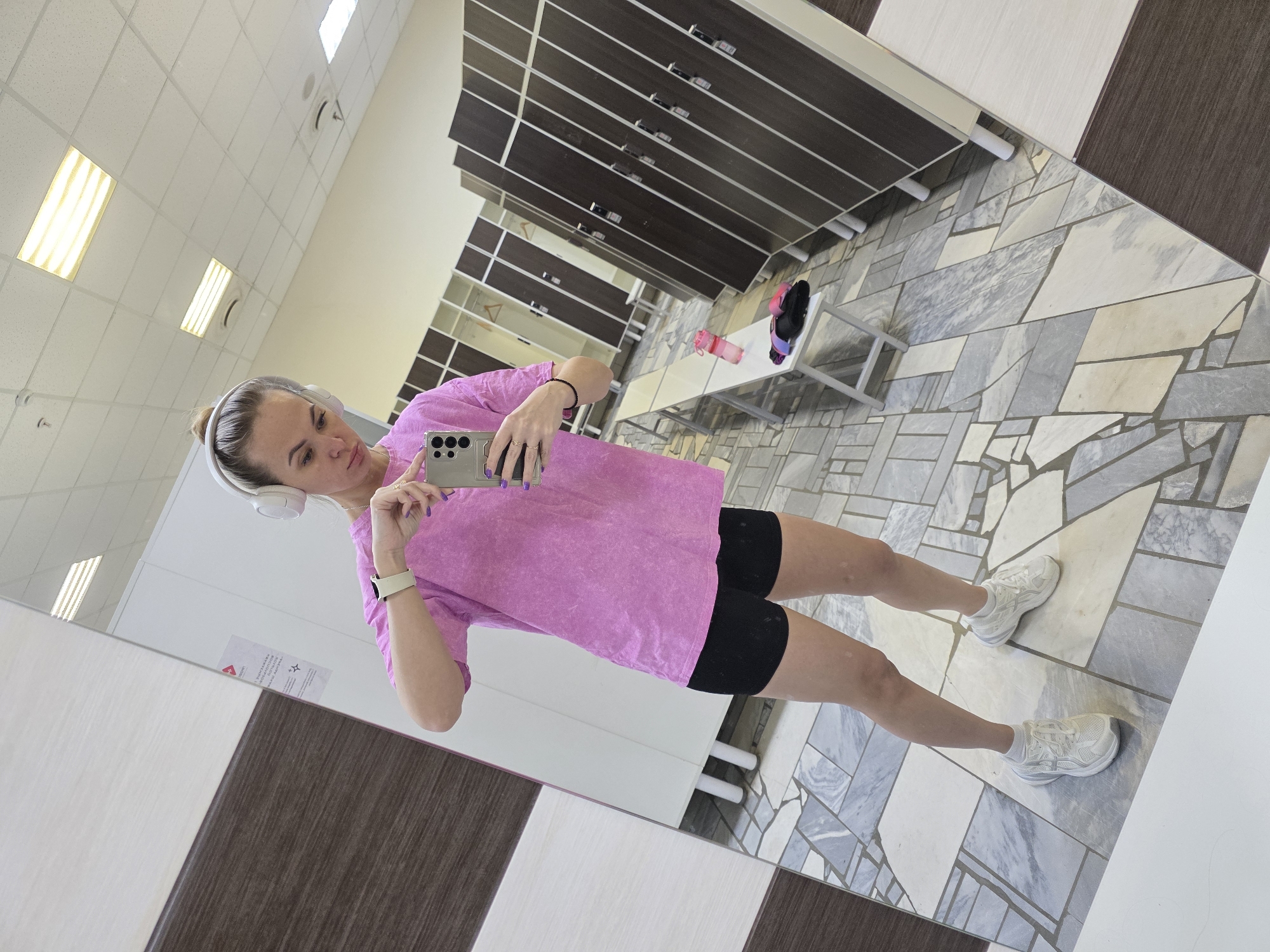 A woman is taking a selfie in a gym locker room. She is wearing a pink t-shirt, black shorts, and white sneakers. She has headphones on and is holding a smartphone in her right hand. The locker room has brown lockers, a gray tiled floor, and a bench. The locker room is located in Chaikovsky, Russia.