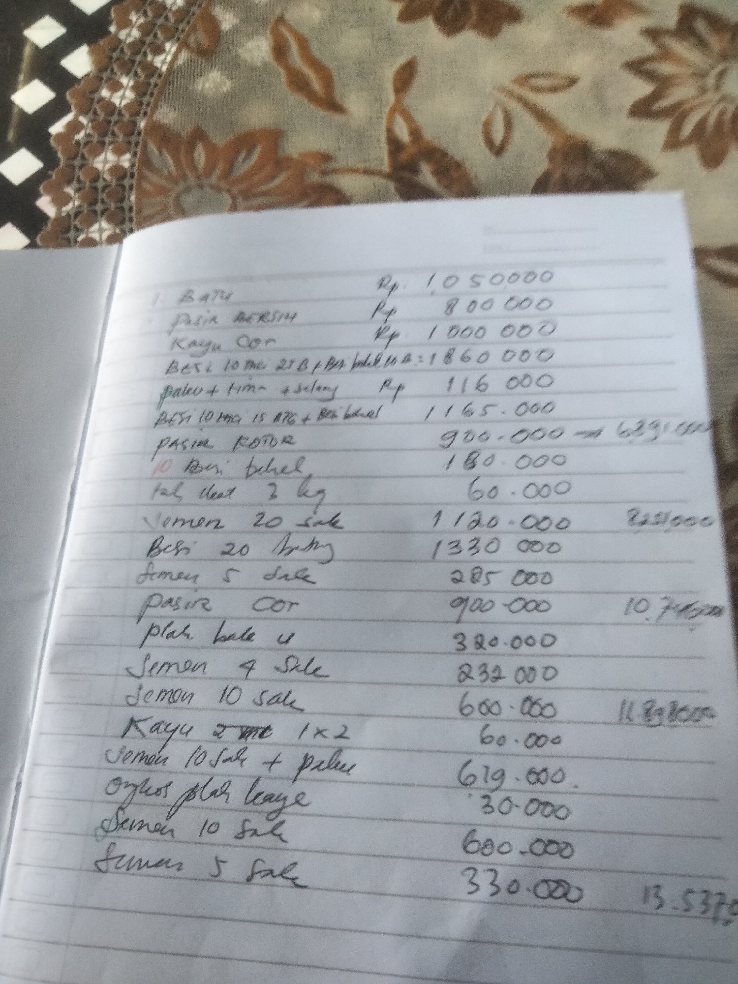 The image shows a handwritten list in a notebook. The notebook is open and lying on a surface with a patterned covering. The list appears to be an itemized bill or record of purchases, with quantities and costs next to each item. The costs are marked with "Rp," which typically denotes Indonesian Rupiah. Some items on the list include building materials such as "Batu" (stone), "Pasir" (sand), "Kayu" (wood), "Besi" (iron), "Semen" (cement), and other related items. Some quantities and descriptions include "20 sak" (20 sacks), "5 sale," and size measurements like "1x2" for lumber. Some totals or calculations are written to the right of the itemized list. The city is not mentioned in this image.
