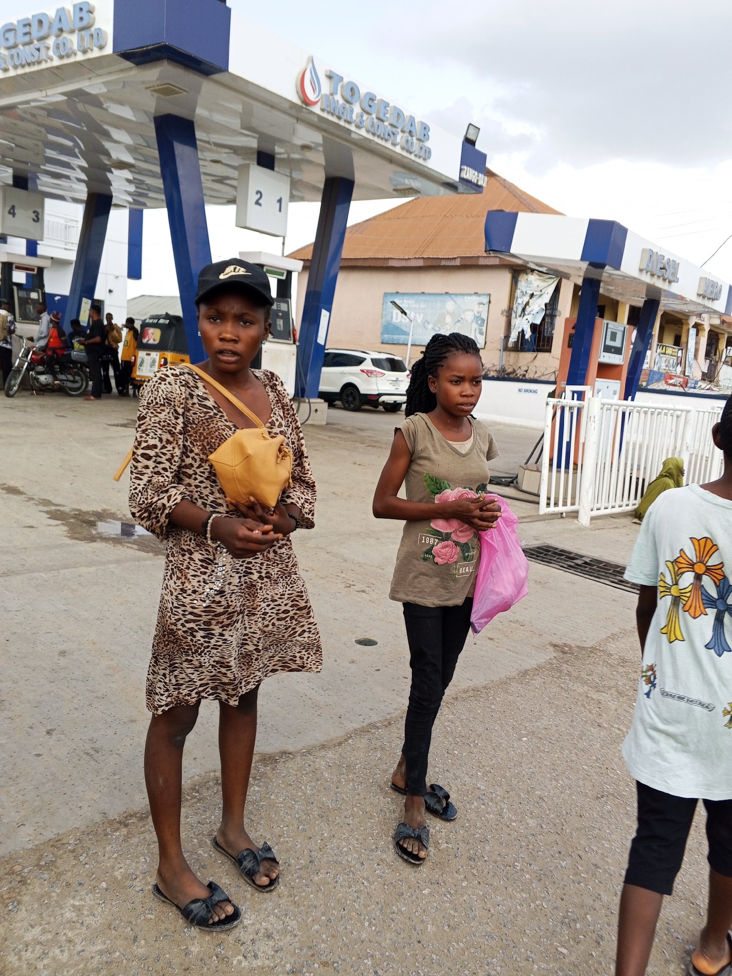 This media file captures a daytime scene at a TOGEDAB gas station in Zango-ndaji, Nigeria, under an overcast sky.
In the foreground, a young woman stands centrally, wearing a leopard-print dress, a black cap with a white logo (possibly Nike), and black sandals. She holds a small tan shoulder bag and looks directly at the camera with a neutral expression. To her right, a younger girl, dressed in a brown t-shirt featuring "1987 BEAU" text and a pink floral design, black pants, and black sandals, holds a pink plastic bag and gazes towards her left. The back of a child wearing a white t-shirt with colorful cross patterns is partially visible on the far right.
The background reveals the gas station forecourt with its distinctive blue and white canopy structure. Visible text on the canopy includes "TOGEDAB OIL & CONST CO., LTD" and "GEDAB CONST. CO., LTD", along with "DIESEL". Fuel pump numbers "2", "1", "4", and "3" are displayed. A "NO SMOKING" sign is mounted on a wall. Several individuals are seen near the pumps, and vehicles such as a red motorcycle, a white SUV, and a yellow tricycle (keke-napep) are present, suggesting routine activities at a fueling station. Buildings with brown roofs are visible behind the station. The ground is paved with concrete. The overall impression is one of everyday life in an urban or semi-urban setting.
