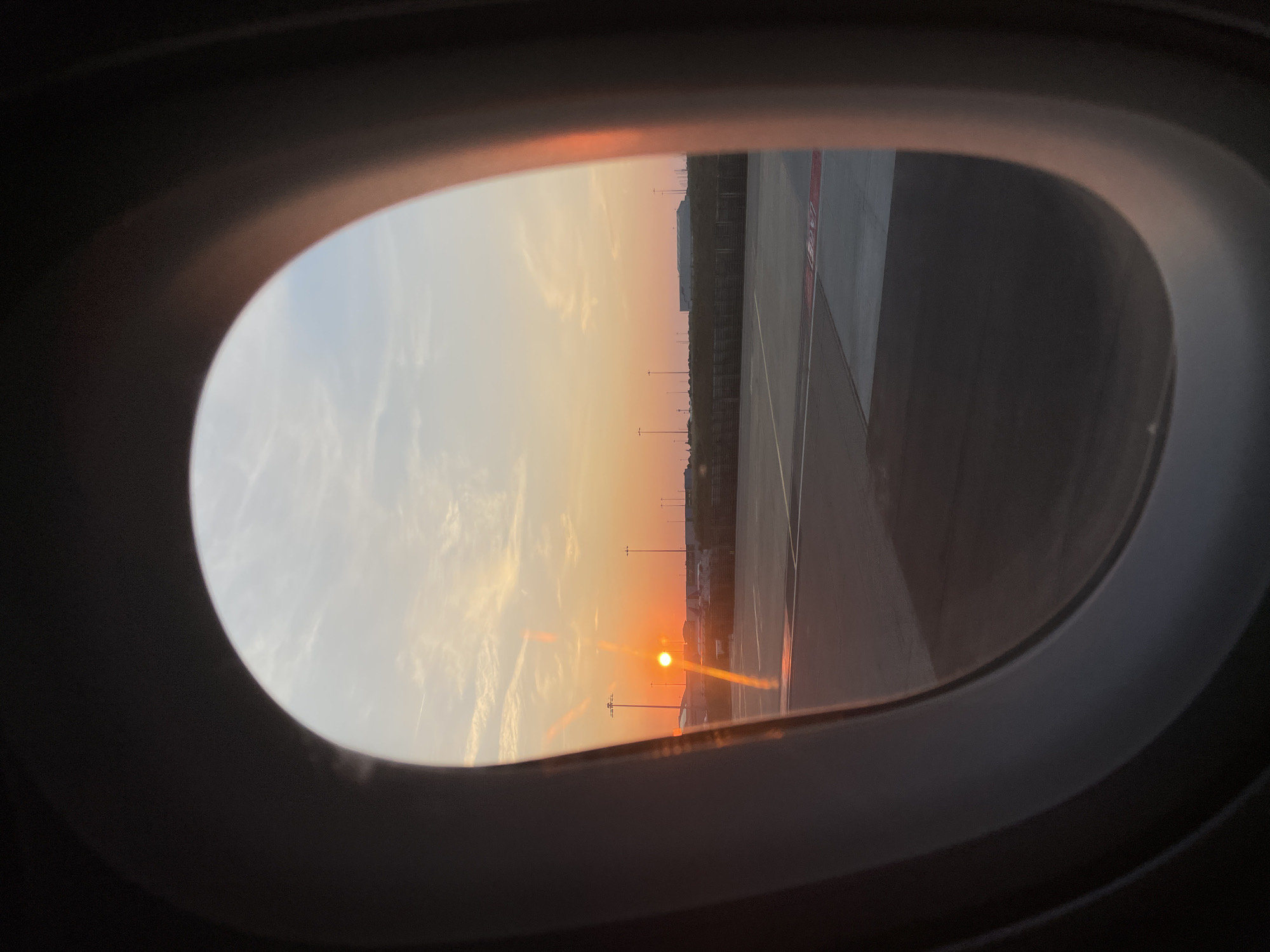 The image shows a view from an airplane window looking out at an airport tarmac during sunset. The sun is low on the horizon, casting a warm orange glow across the sky and reflecting on the tarmac. Several lampposts and airport buildings are visible in the distance. The edge of the airplane window frame is visible in the foreground, obscuring parts of the view. This image was taken at London Luton Airport, United Kingdom.