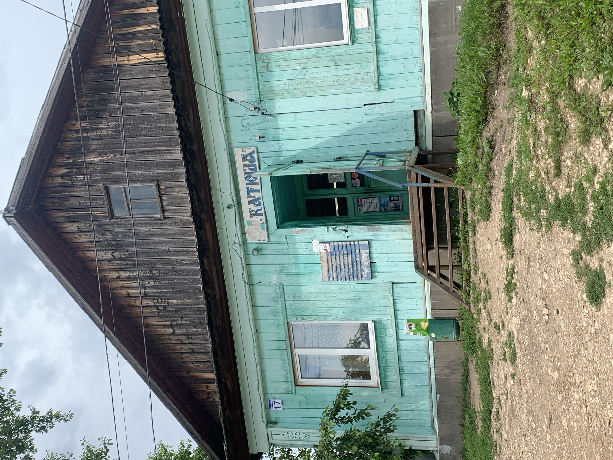 The image shows the exterior of a weathered, turquoise-painted wooden building with a dark, aged wooden gable. The building appears to be a shop, as indicated by a sign above the entrance that reads "KATIOXA" in Cyrillic letters. The entrance is an open doorway with a blue railing leading up to it. There is a window to the left of the entrance with white framing and a patterned curtain visible inside. Another window with white framing is located to the right of the entrance. A blue and white sign with Cyrillic text is affixed to the wall to the left of the entrance, and a similar, partially obscured sign is on the wall to the right of the doorway. A green trash receptacle with a white and yellow wrapper on top is situated to the left of the entrance stairs. The ground in front of the building is covered in dirt and sparse green vegetation. The building is situated in what appears to be a rural or suburban area, with power lines running across the facade and a tree with green leaves on the left side of the frame. The sky above is overcast with gray clouds. The address "17" is visible on a blue and white sign to the left of the main entrance. The context of the city of Барсаи, Russia is not visually derivable from the image itself.