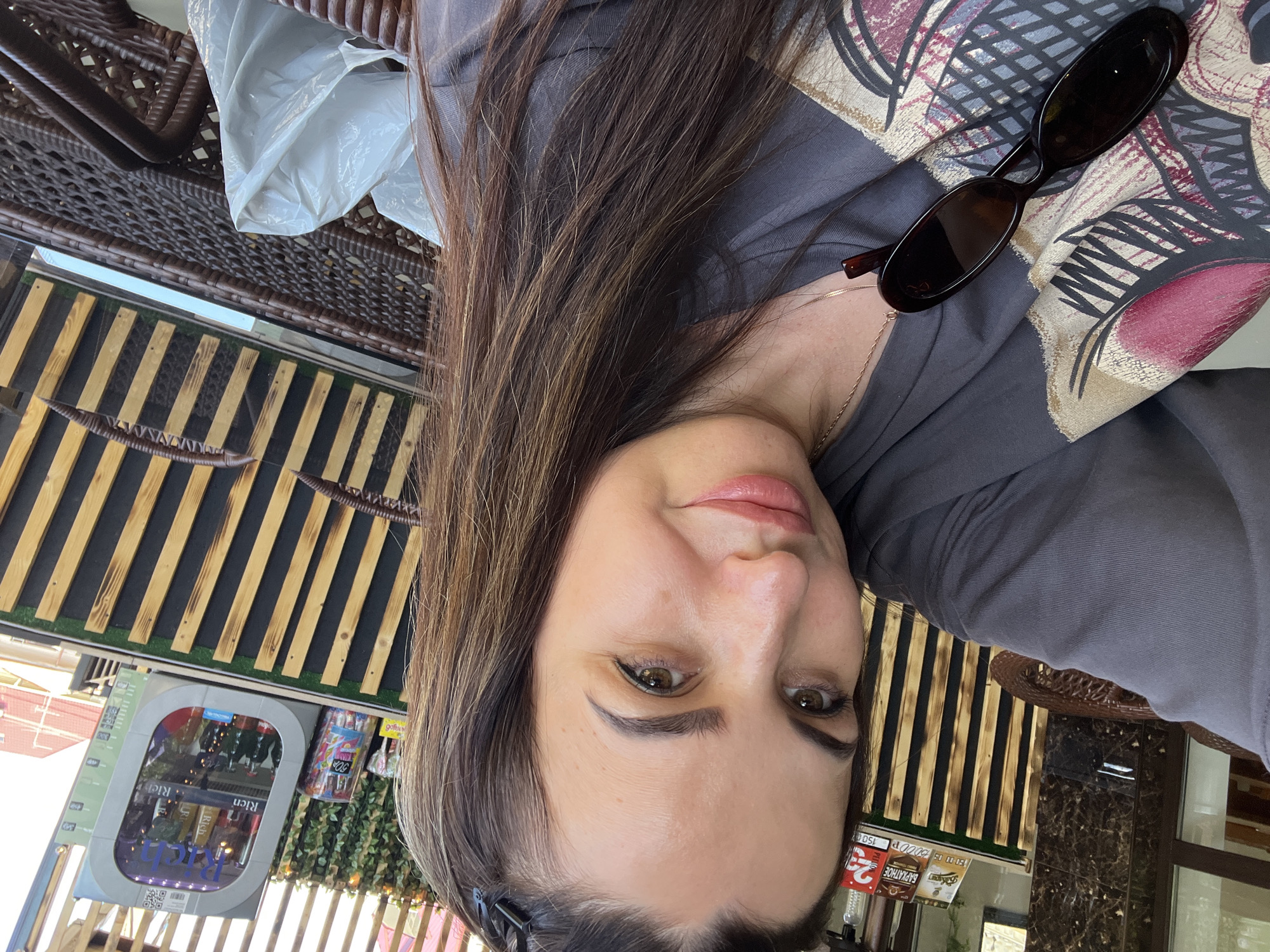 A woman is taking a selfie outdoors. She has long, brown hair and is wearing a gray t-shirt with a graphic design on the front. She is also wearing sunglasses on her shirt. In the background, there is a display of drinks in a cooler, a wooden fence, and some plants. The scene appears to be a cafe or outdoor seating area.