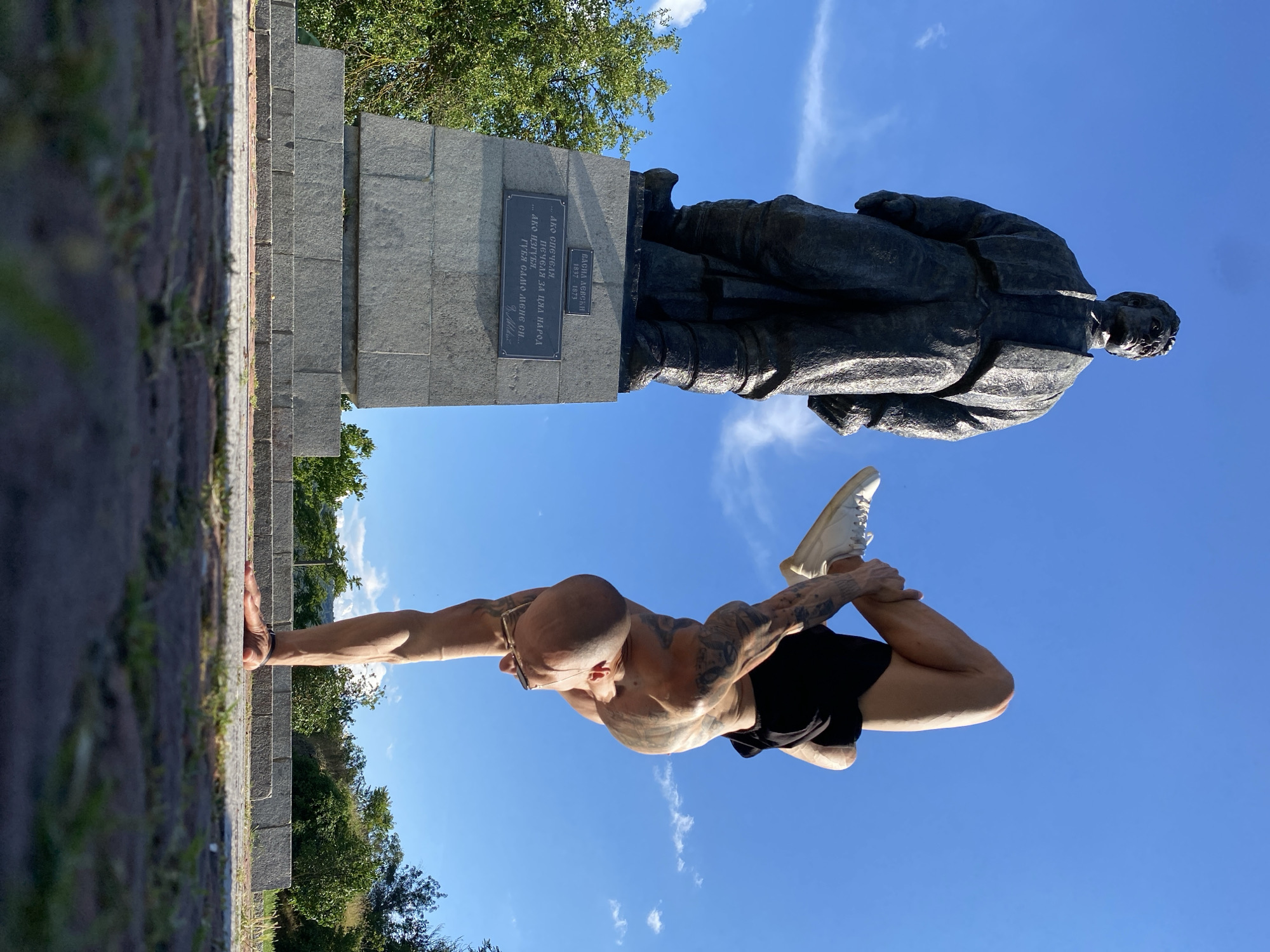 An athletic man with visible tattoos performs a handstand with his body contorted in front of a bronze statue on a sunny day. The statue, located in the city of Lovech, Bulgaria, depicts a historical figure and stands on a granite pedestal with an inscription. The man is wearing black shorts and white sneakers. The background consists of a clear blue sky with scattered clouds and lush green trees. The ground is paved with stone.