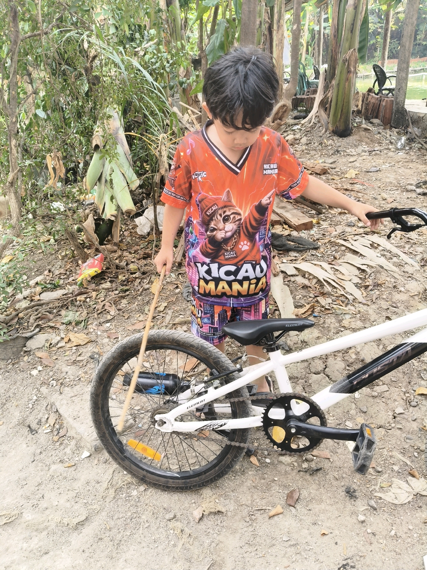 A young boy stands next to a white BMX bicycle, holding a stick. He is wearing an orange t-shirt with a graphic of a cat and the words "KICAUMANIA" and shorts with a similar design. The bicycle has a black seat, black pedals, and black handlebars. The boy is looking down at the bicycle. The scene appears to be outdoors in an unpaved area with dirt and leaves on the ground. In the background, there are trees and some debris. The location is identified as South Tangerang, Indonesia.