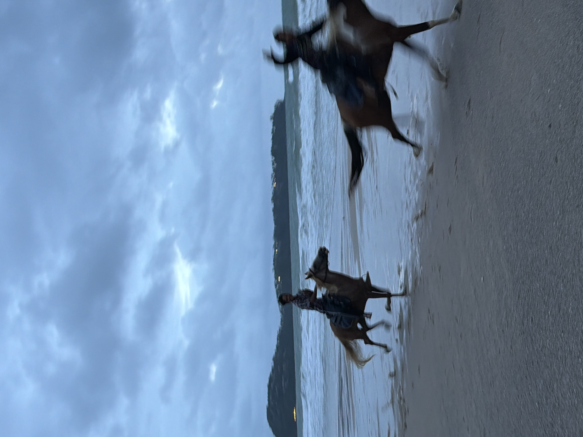 The photograph captures two blurry figures on horseback riding along a beach. The horses are partially submerged in the water near the shoreline, and the sky is overcast with heavy clouds. The scene takes place on a beach near Choeng Thale, Thailand, as evidenced by the background's landscape, which features a forested area with a few lights visible along the distant shoreline.