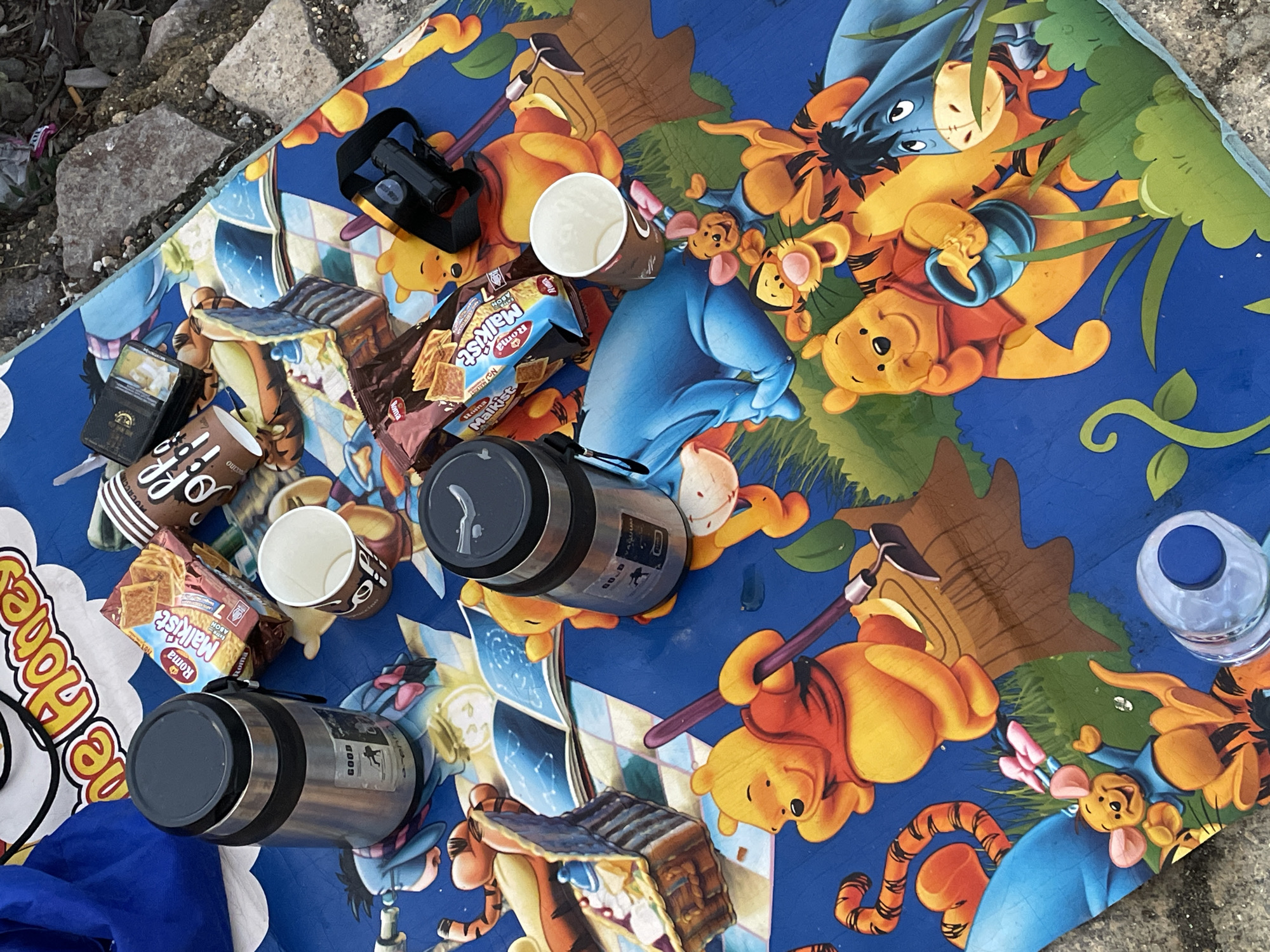 The image shows a picnic scene on a Winnie the Pooh-themed picnic blanket, possibly in Unknown, Indonesia, based on the "Roma Malkist" crackers, which are an Indonesian brand. The blanket is spread on what appears to be a stone or concrete surface.
Several items are scattered across the blanket:
* Two stainless steel vacuum flasks.
* Several disposable coffee cups, some of which are empty.
* Two packets of "Roma Malkist" brand crackers, one of which is open.
* An open box of cookies.
* A black headlamp.
* A cigarette lighter.
* An empty plastic bottle.
The blanket features various Winnie the Pooh characters. The overall impression is that someone has recently had a picnic.