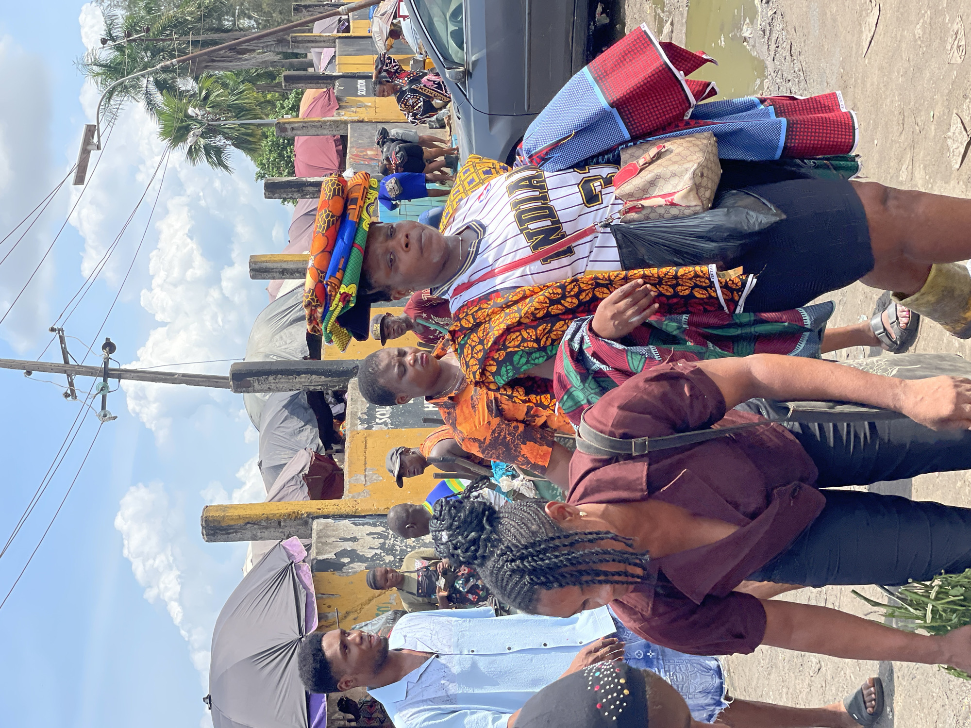 This image captures a bustling outdoor scene, likely a market or a busy street, under bright daylight with a partly cloudy sky. The photograph is oriented sideways (rotated 90 degrees clockwise).
In the foreground, a woman is prominently featured, looking directly at the viewer. She carries a stack of vibrantly patterned fabrics on her head and another bundle of colorful textiles draped over her arm and shoulder. She is wearing a white and black striped baseball-style jersey with "INDIA" visible across the chest, over a patterned garment. A patterned handbag and a black plastic bag are slung over her shoulder. Behind her, another woman also carries patterned fabrics.
Several other individuals are scattered throughout the mid-ground and background, indicating a crowded environment. Some appear to be walking, while others might be vendors or shoppers. A man in a light blue shirt stands in the lower left portion of the image, facing right. In the lower right, a person with braided hair, wearing a maroon top, is visible.
The setting includes utility poles with wires stretching across the sky, suggesting an urban or semi-urban area. Palm trees and other green foliage are visible in the distance. Concrete barriers or structures, some painted yellow, line the ground. A dark-colored car is partially visible in the mid-ground. Several umbrellas, either closed or partially open, are seen, possibly providing shade for vendors or pedestrians. The ground is uneven, with patches of dirt and what appears to be a puddle or muddy area on the far right.
The visible text includes "INDIA" on the main subject's jersey and "SOLUTION" on a yellow structure in the background. The overall activity suggests commercial exchange or daily movement through a vibrant public space.
Location context: Unknown, Unknown.