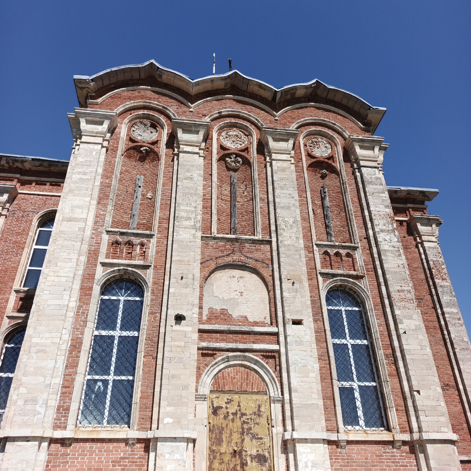 This is a ground-level, slightly low-angle, full shot of the exterior of an old, weathered building, likely a public institution, located in the city of Tugustemire, Russia. The building is constructed primarily of red brick with contrasting lighter stone elements. The sky is a clear, vibrant blue with no clouds.
The architectural style appears to be a blend of Neo-Gothic and possibly some other European influences, with arched windows, decorative brickwork, and stone detailing. The facade is asymmetrical and features multiple stories.
On the left side of the building, there are two tall, narrow windows with white frames and what appear to be decorative metal grates. The lower window is partially obscured by the angle of the shot.
The central section of the building is dominated by a large, arched entryway. The door itself is made of heavily rusted metal, with peeling paint revealing a yellowish-brown undercoat. Above the doorway, there's a large, empty arched recess, also made of brick.
Flanking the central section are prominent vertical stone columns. Between these columns and above the arched recess, there are several smaller, arched niches. Some of these niches contain what appear to be remnants of decorative stonework, while others are empty or contain weathered, possibly wooden, elements. In one of these niches, a vertical piece of rough wood is visible, possibly a structural remnant or a piece of old ornamentation.
The upper part of the building features a decorative roofline with a scalloped or wavy edge, topped with a stone coping. On the roof, two slender white poles are visible, along with a dark, angled object, which could be part of an antenna or a weather vane.
The building shows signs of age and neglect, with chipped paint, weathered brickwork, and areas of crumbling plaster. Despite its condition, the building conveys a sense of historical significance and architectural grandeur. There are no people visible in the image. The environment appears to be an outdoor setting, with the building as the sole subject of focus.