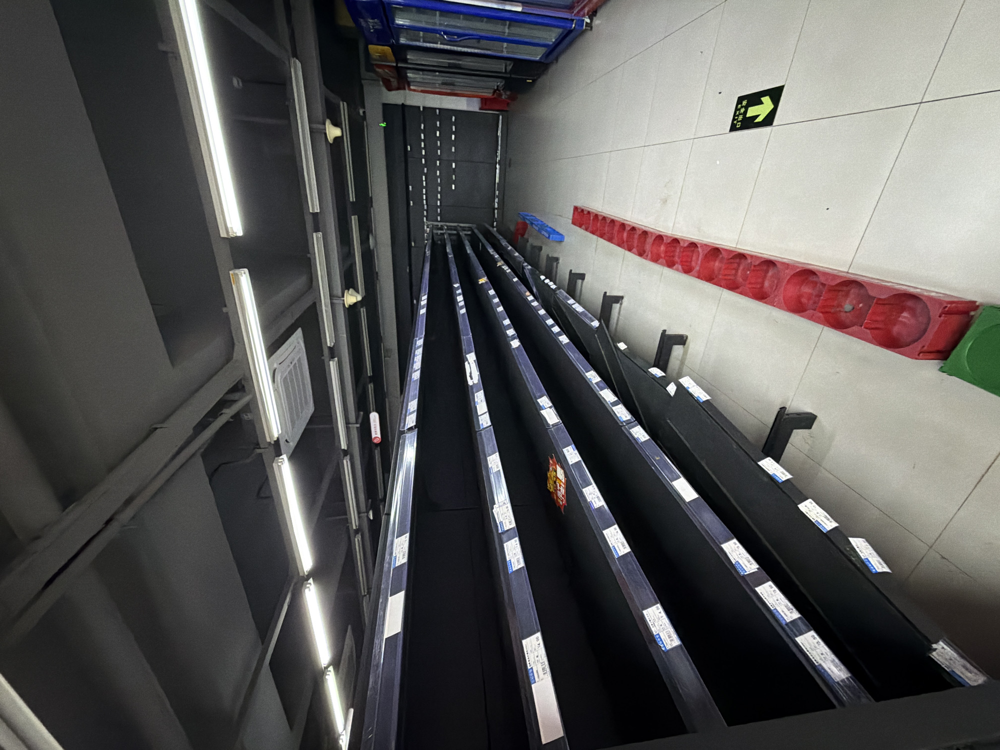 The image depicts the interior of a supermarket in Jiangjin, China. The majority of the shelves are empty, suggesting a lack of goods or a recent restocking event. The lighting is provided by fluorescent fixtures on the dark ceiling. In the foreground, there are rows of empty black shelves. On the floor, there is a red display rack designed to hold circular items, and a green recycling bin. Further back, there are refrigerators, and in the distance, a closed roll-up door. A yellow directional sign with Chinese characters is visible on the tiled floor pointing to an exit. No people are present in the image.