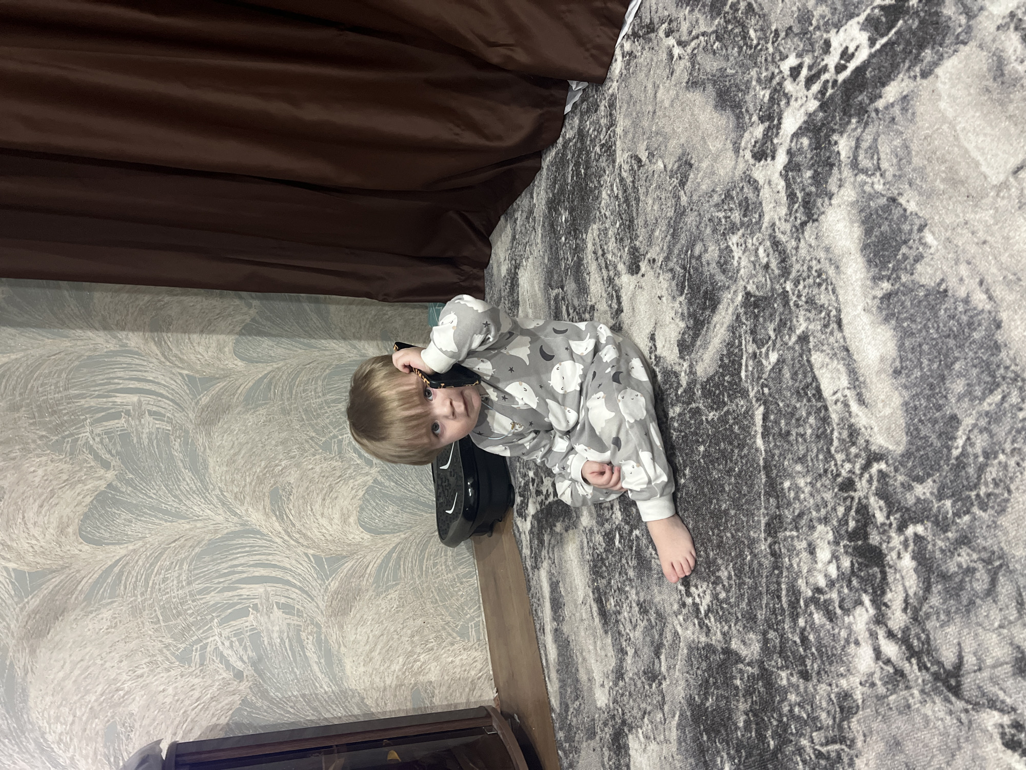 This indoor scene, located in Noviy, Russia, features a young child seated on a multi-toned grey and white patterned rug. The child has light-colored hair and is dressed in grey pajamas with a repeating white pattern of ghosts and crescent moons. Their left foot, bare, rests on the rug. The child holds a dark-colored object, resembling a phone or toy phone, to their right ear with their right hand, facing forward with a neutral expression. In the background, to the child's left, a dark wooden furniture piece is partially visible. Behind the child, a black robotic vacuum cleaner or similar device is positioned on the rug. The wall behind the child is covered with light blue-grey wallpaper featuring a white, feathery or abstract foliage design. To the right side of the frame, a plain, dark brown curtain extends from the top to the bottom, partially obscuring the background.