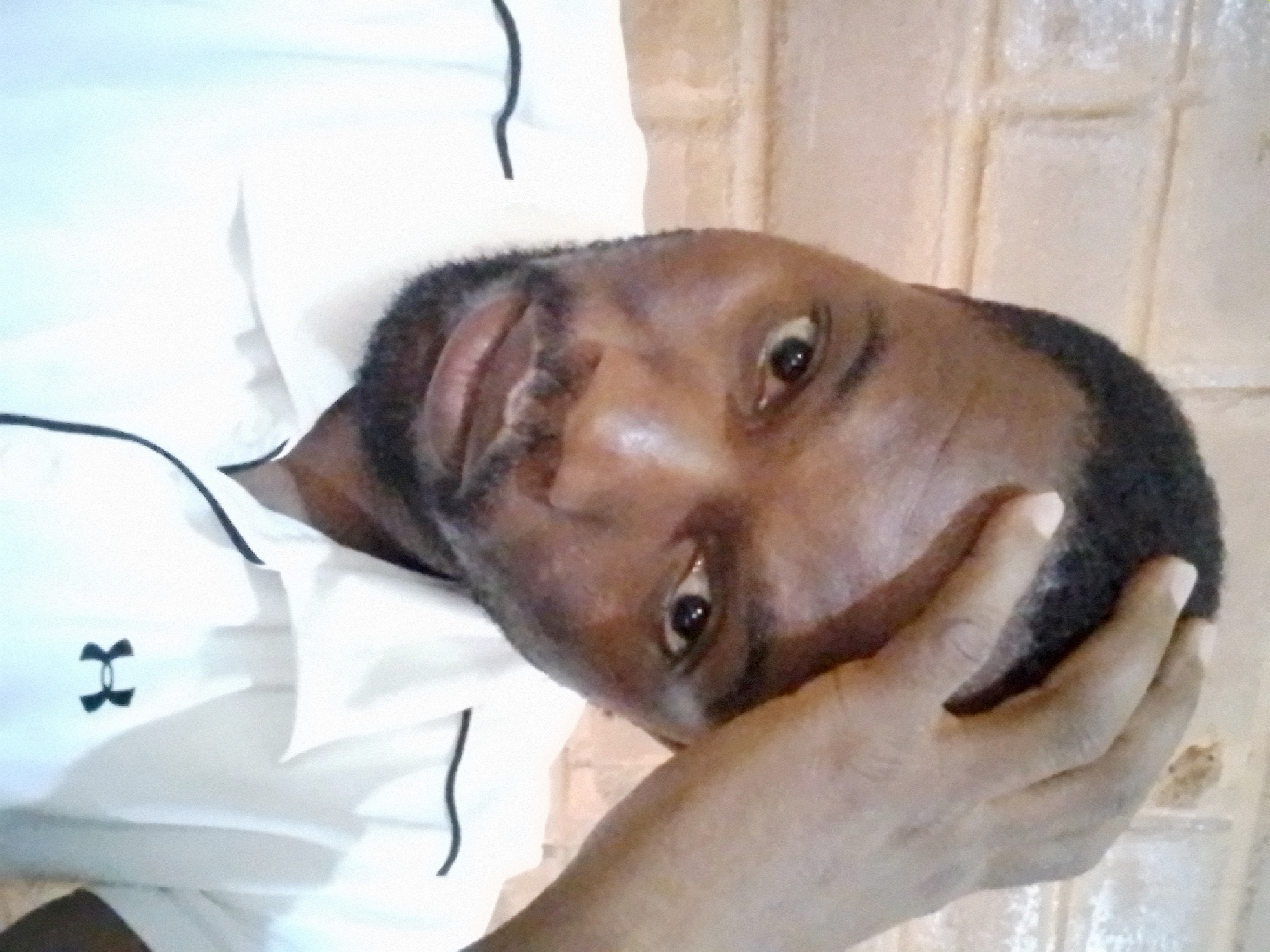 A man is captured in a selfie, his right hand resting on his forehead. He has short, dark hair and a beard. He is wearing a white polo shirt with black trim along the collar and sleeves, and the Under Armour logo is visible on the left side of his chest. The background consists of a brick wall. The image is taken indoors.
